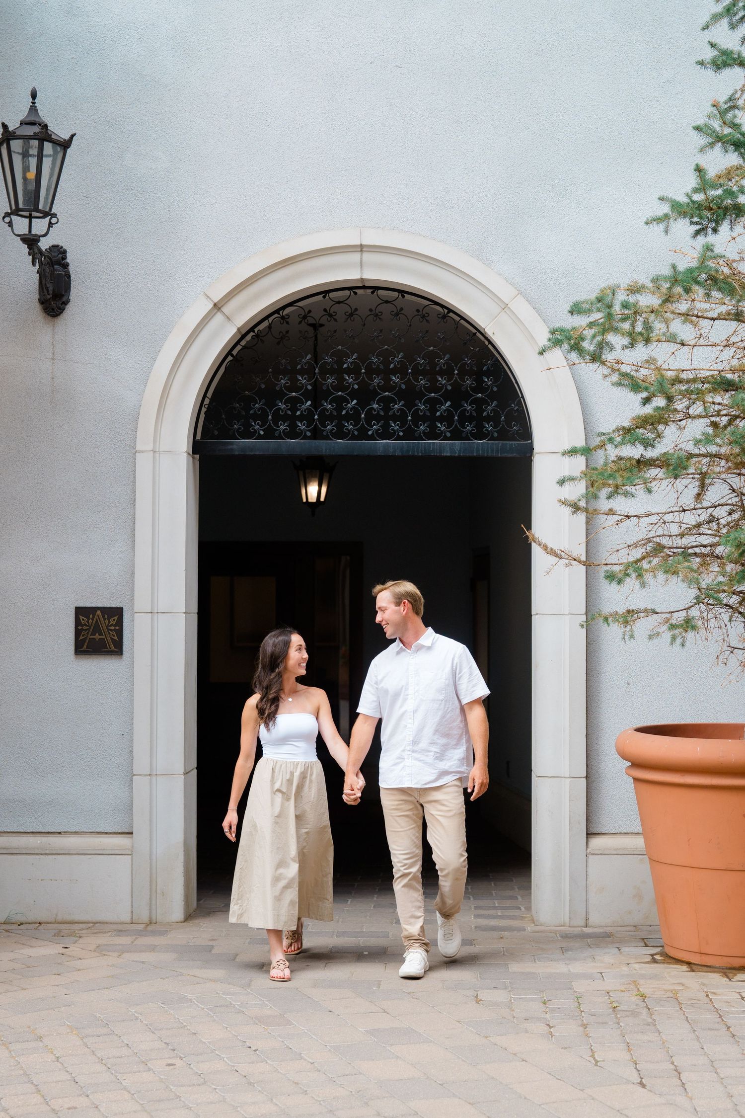A couple walks together through a series of white stone archways while holding hands on a stone patio.