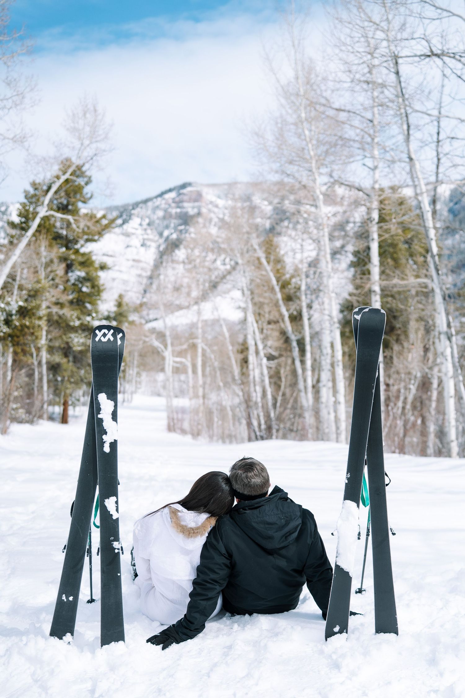 Two people sitting in snow between upright skis on a winter mountain slope with bare trees in the background.