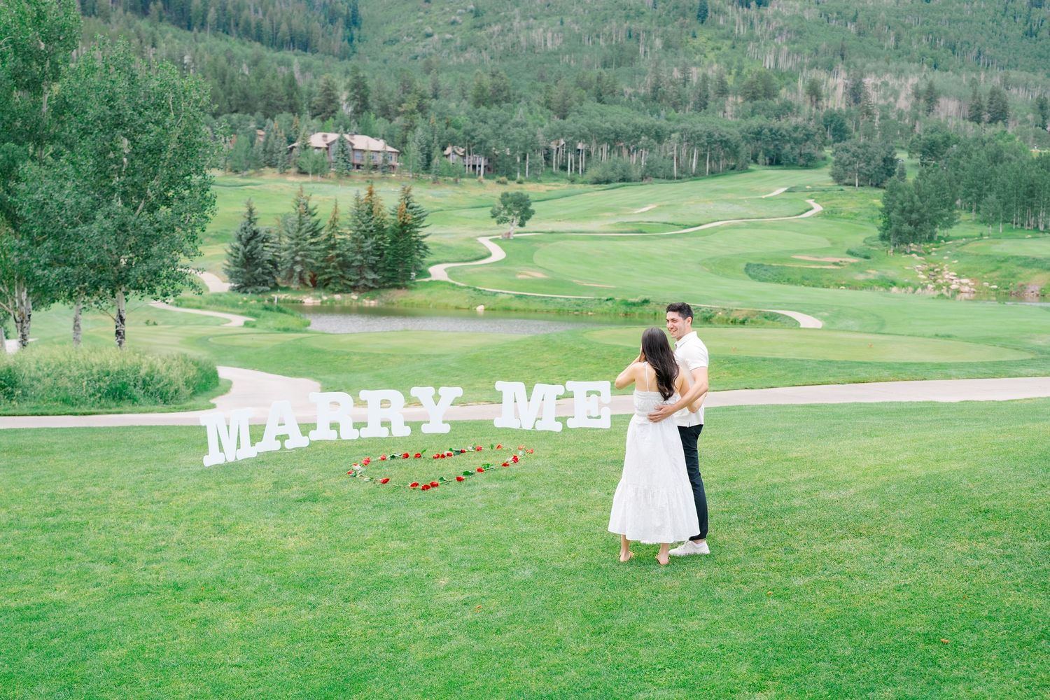 A couple shares a romantic moment on a lush green hillside overlooking a golf course in a mountain setting.