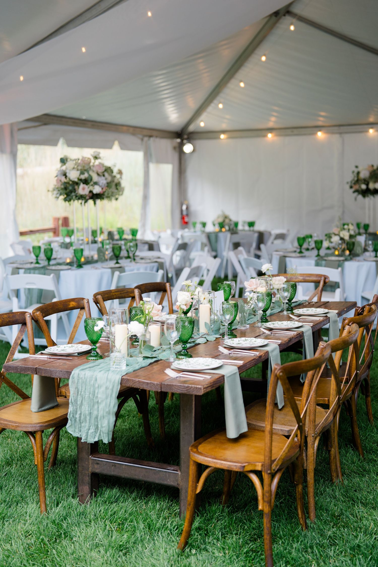Rustic wooden farm tables with green glassware set for an elegant outdoor wedding reception under a white tent.