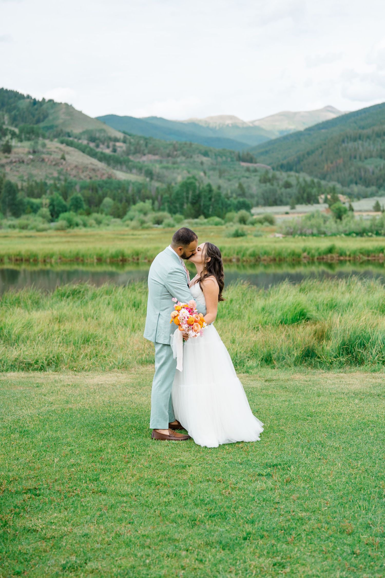 Romantic wedding couple shares intimate moments on a grassy field with mountain scenery.