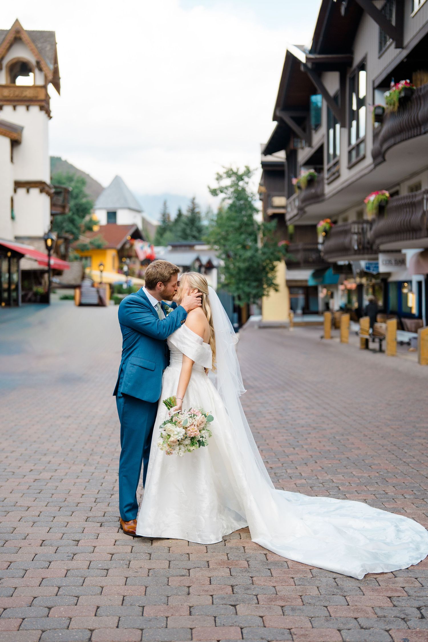 A romantic wedding couple embraces on a charming European-style cobblestone street with traditional buildings in the background.