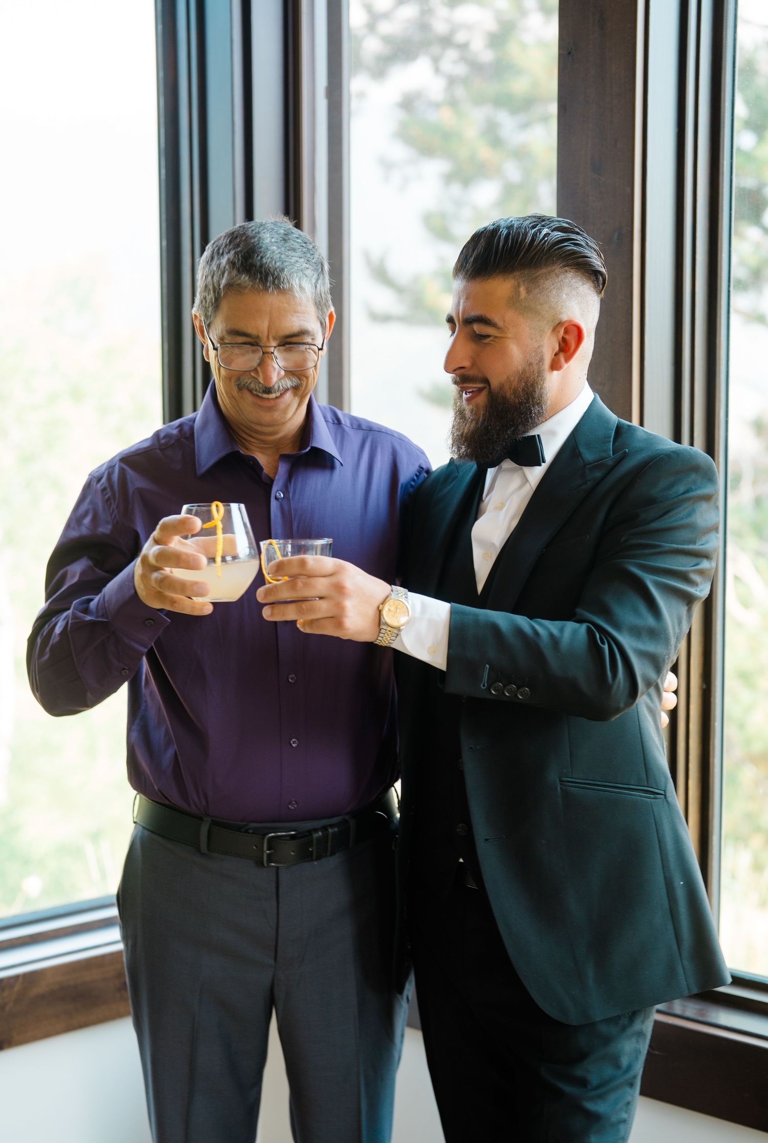 Two people share drinks and laughter together by large windows in formal attire.