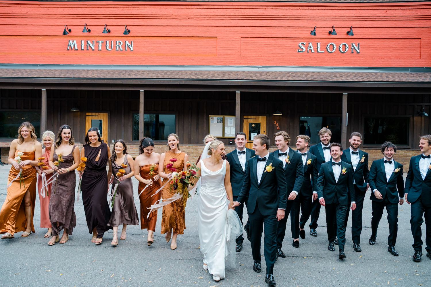 Wedding party lines up against a coral-colored wall wearing formal attire in coordinated colors.