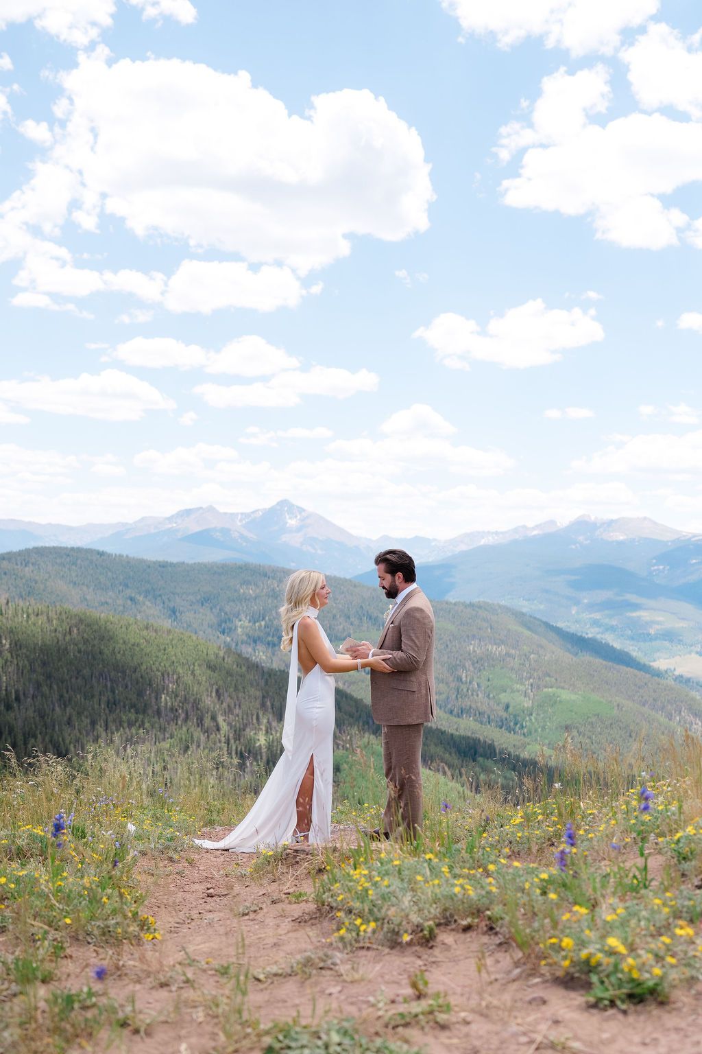 Couple exchanging vows on a mountain summit surrounded by wildflowers and scenic Rocky Mountain views on their wedding day.