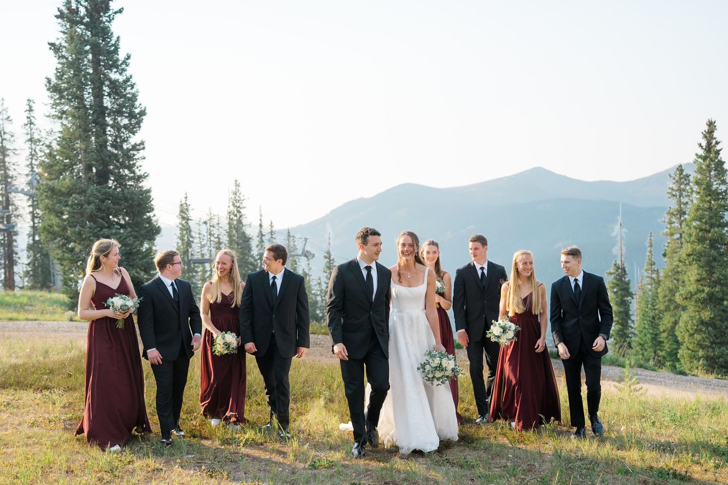 Wedding party walks outdoors in mountain meadow, bridesmaids in burgundy gowns and groomsmen in black suits flanking bride and groom.