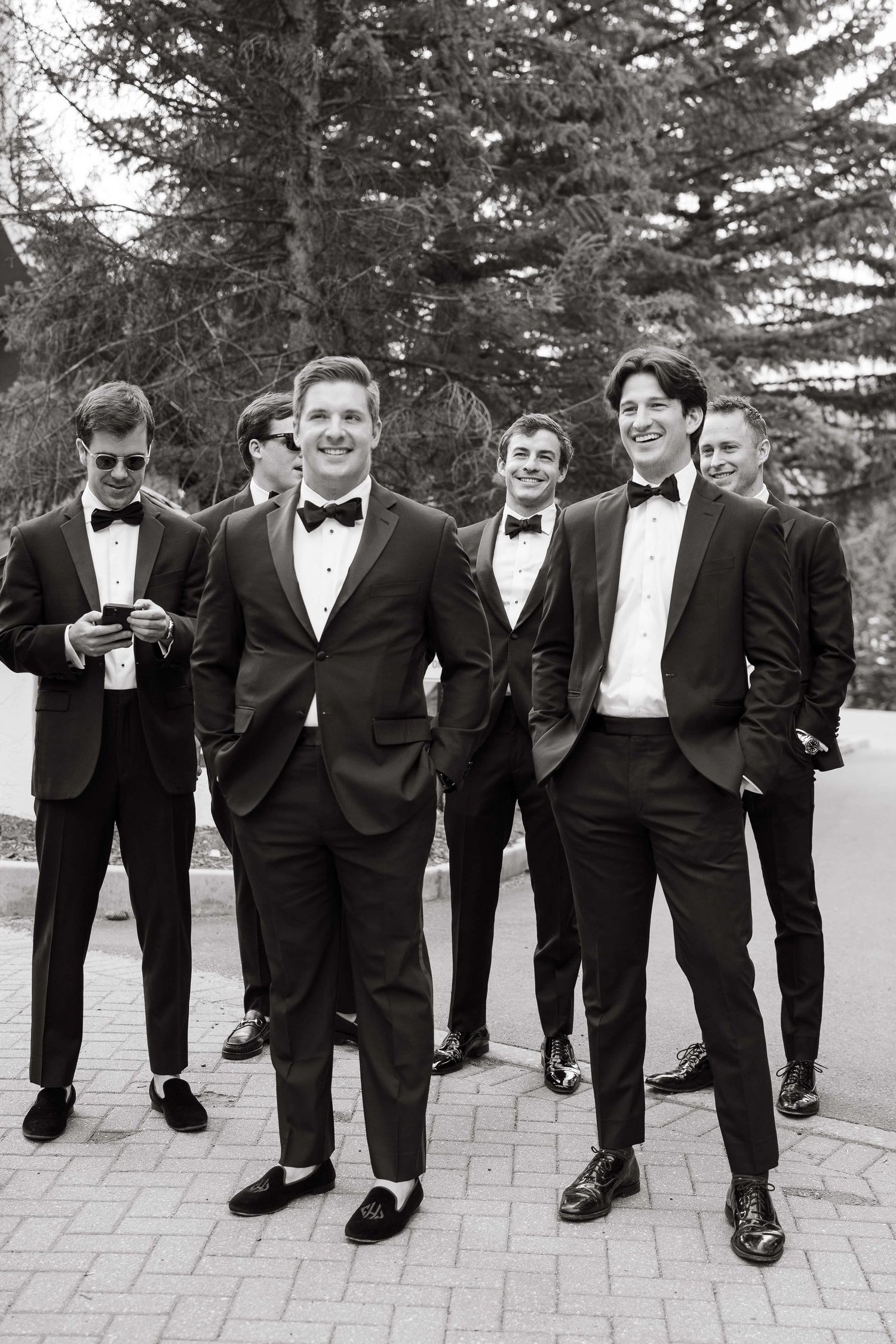 Black and white photo of groomsmen in tuxedos with bow ties posing outdoors near evergreen trees.