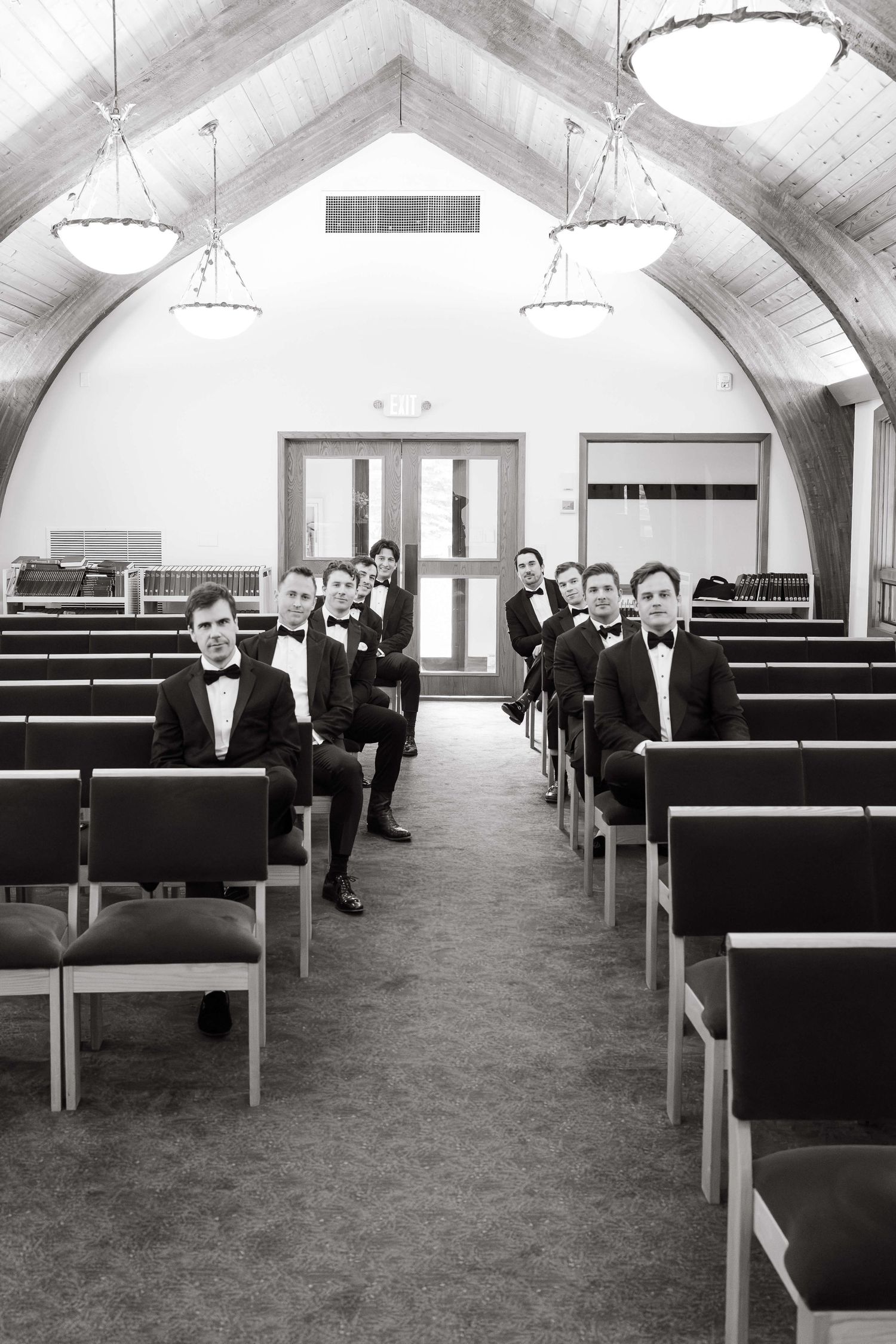Groomsmen in tuxedos pose in church pews along the aisle in a black and white wedding photo.