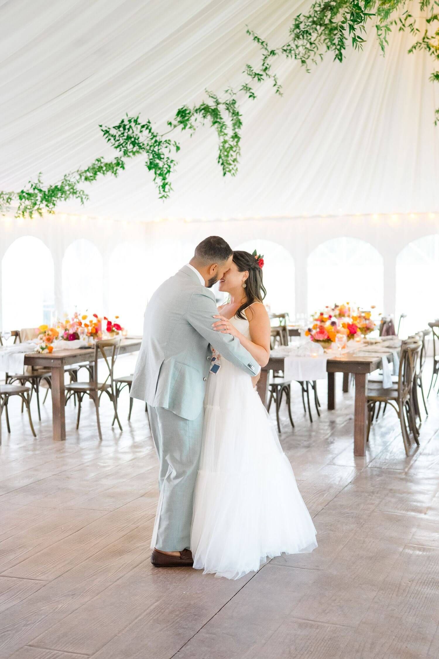 A couple shares an intimate moment during their first dance in an elegant tent reception with greenery decor and rustic tables at Camp Hale
