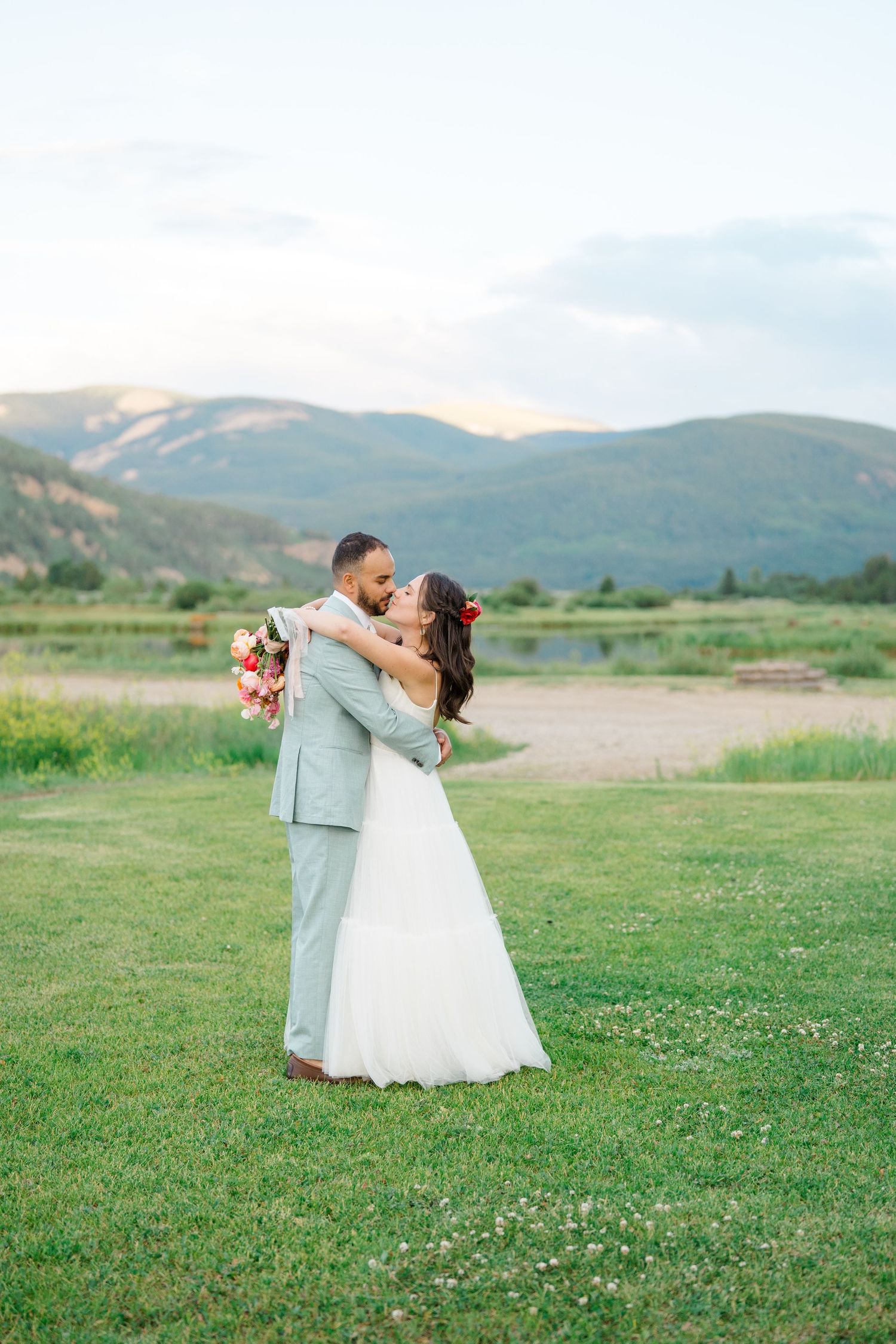 A couple shares a romantic dip kiss in a scenic mountain meadow with green grass and rolling hills in the background.