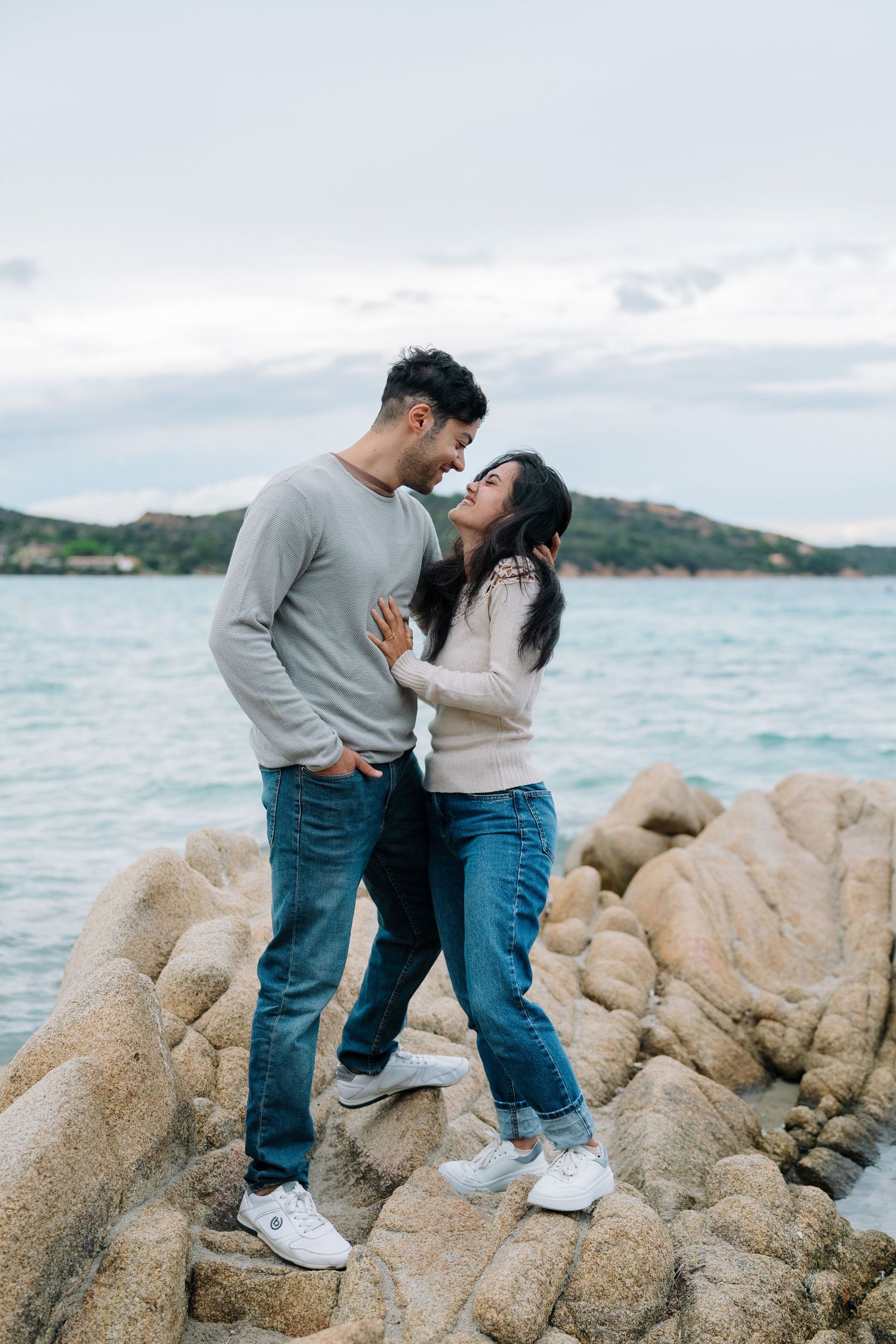A couple stands together on coastal rocks by the ocean during a romantic photoshoot at sunset.