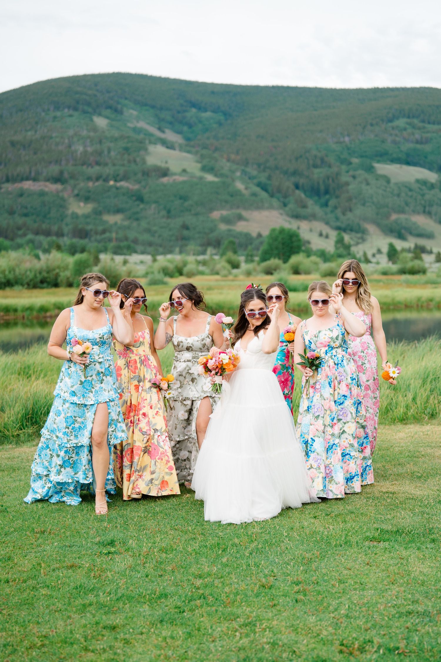 Bridesmaids in bright floral dresses gather on a grassy hill with mountain views.