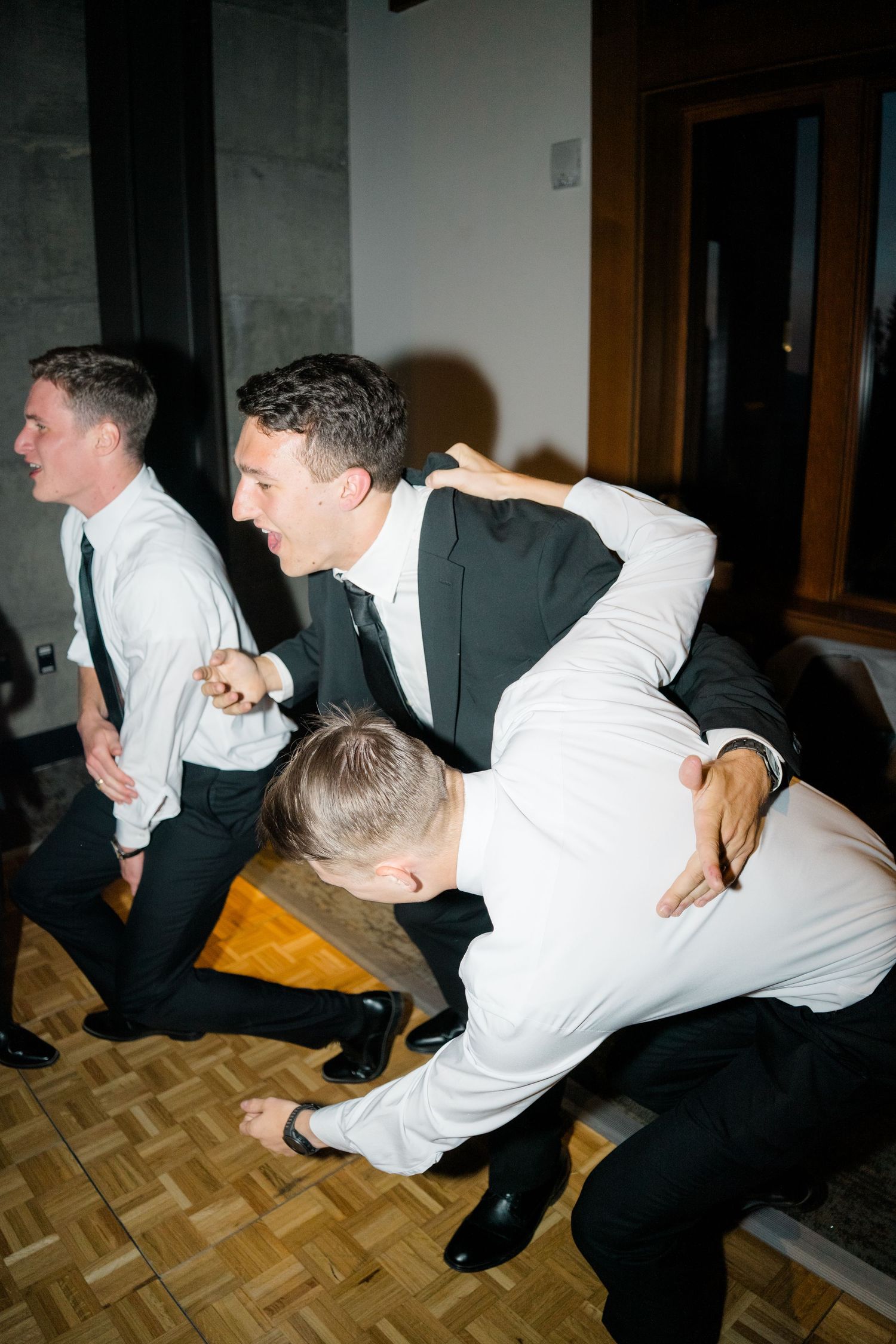 Groomsmen in formal attire playfully dance and pose on a parquet floor during wedding reception.