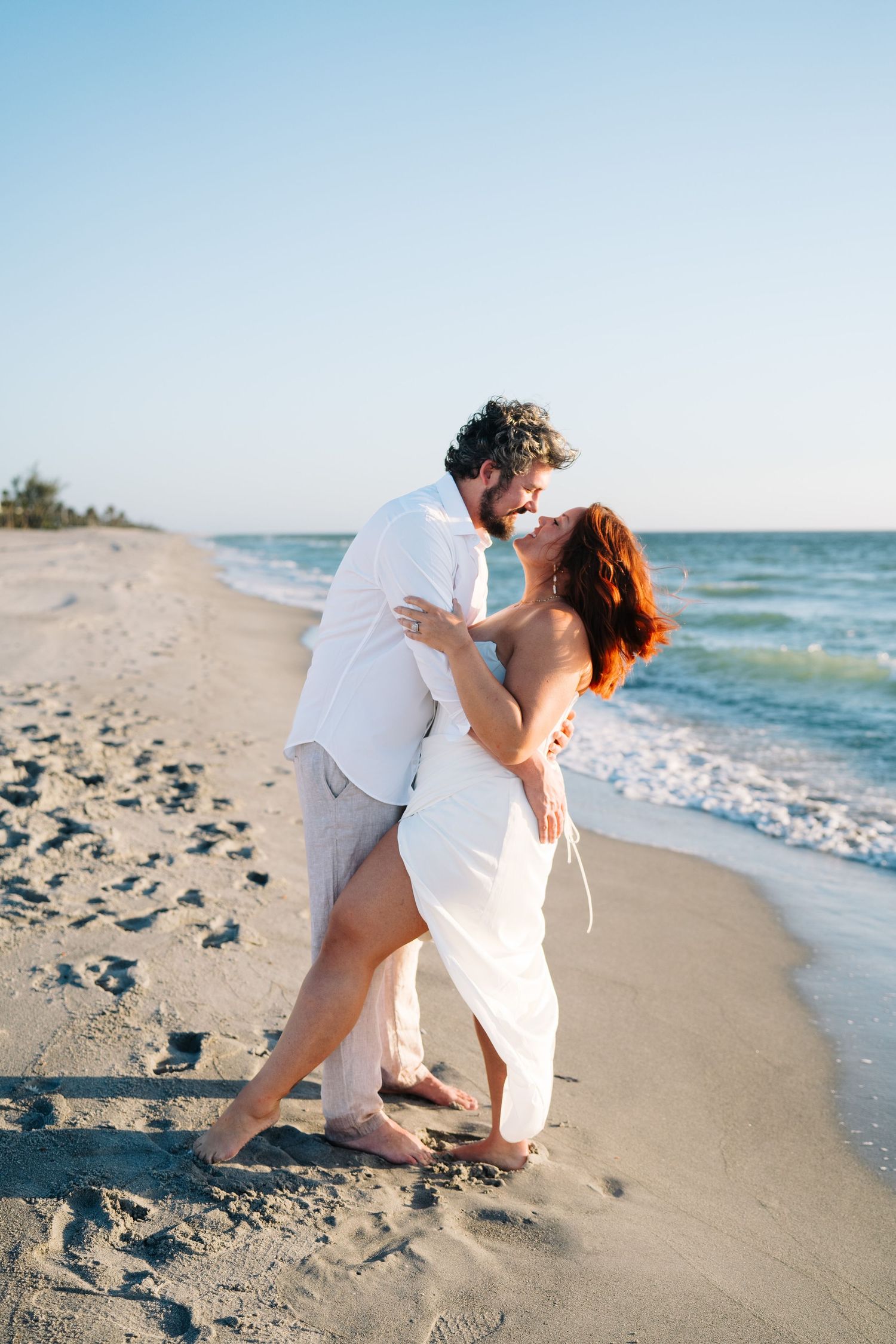 A romantic couple embraces while dancing together on a sunny beach with waves and blue sky in the background.