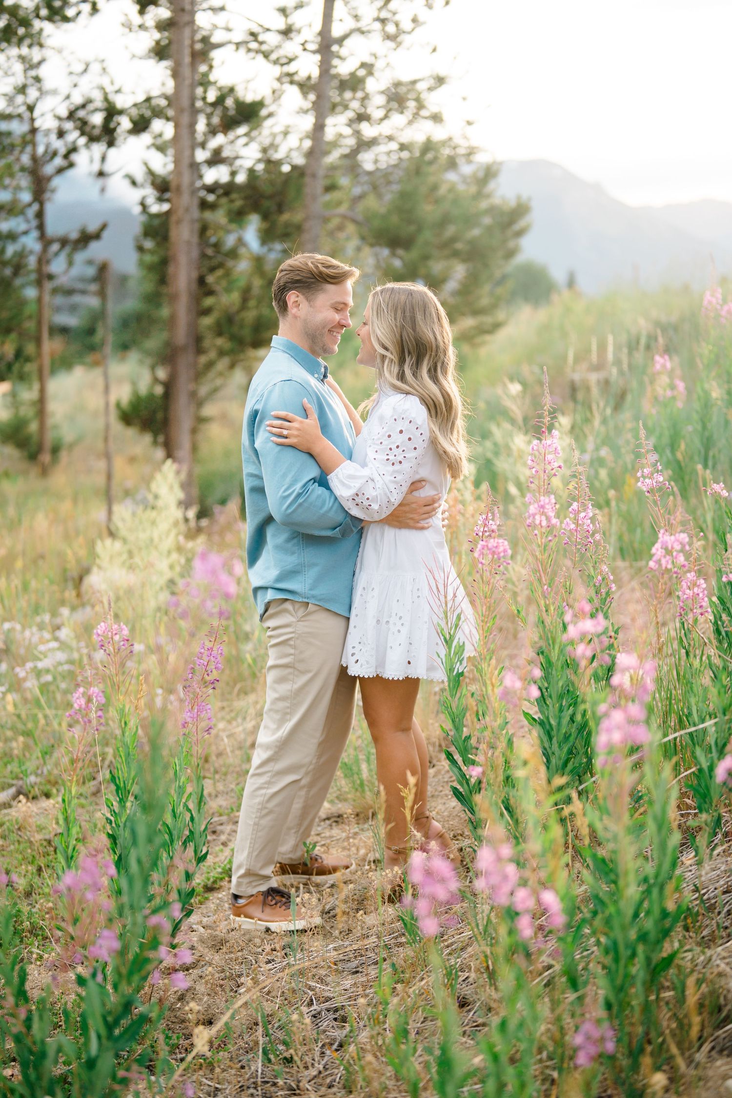 A couple embraces in a mountain meadow filled with blooming pink wildflowers on a sunny summer day.