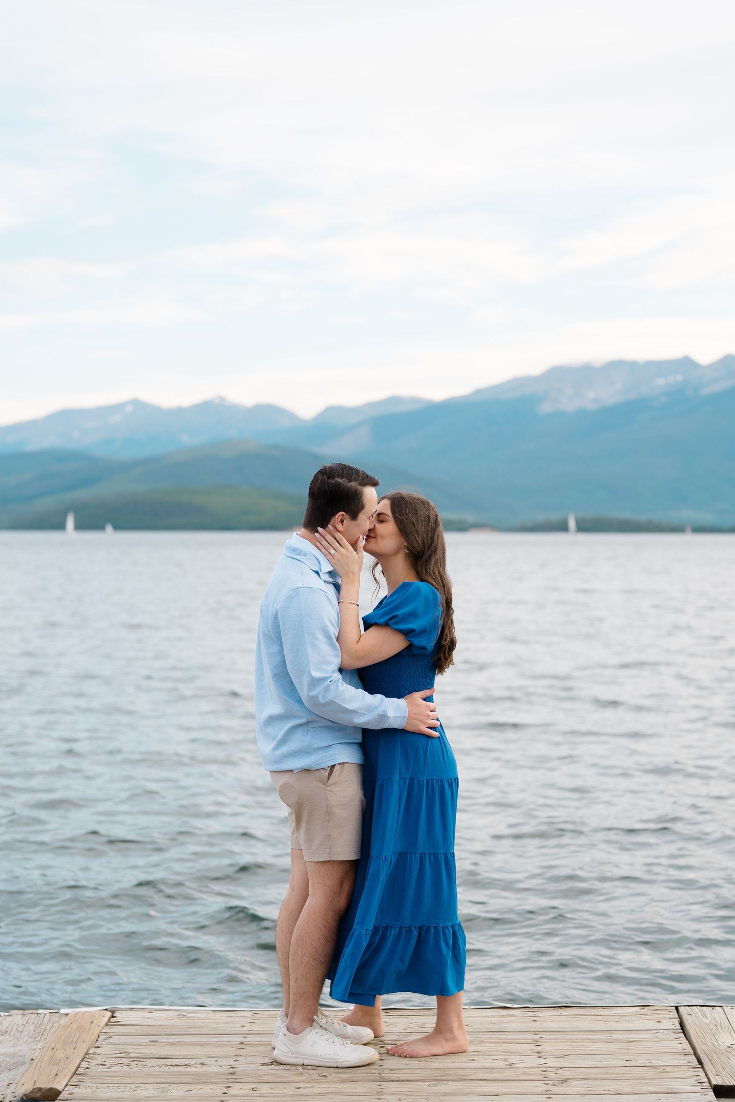 A couple shares a romantic kiss on a wooden dock overlooking a mountain lake with sailboats in the distance.