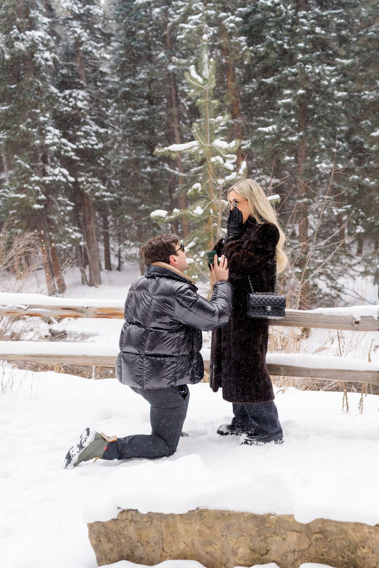 Man proposing to woman in snowy winter forest, kneeling with ring box as she covers her mouth in surprise.