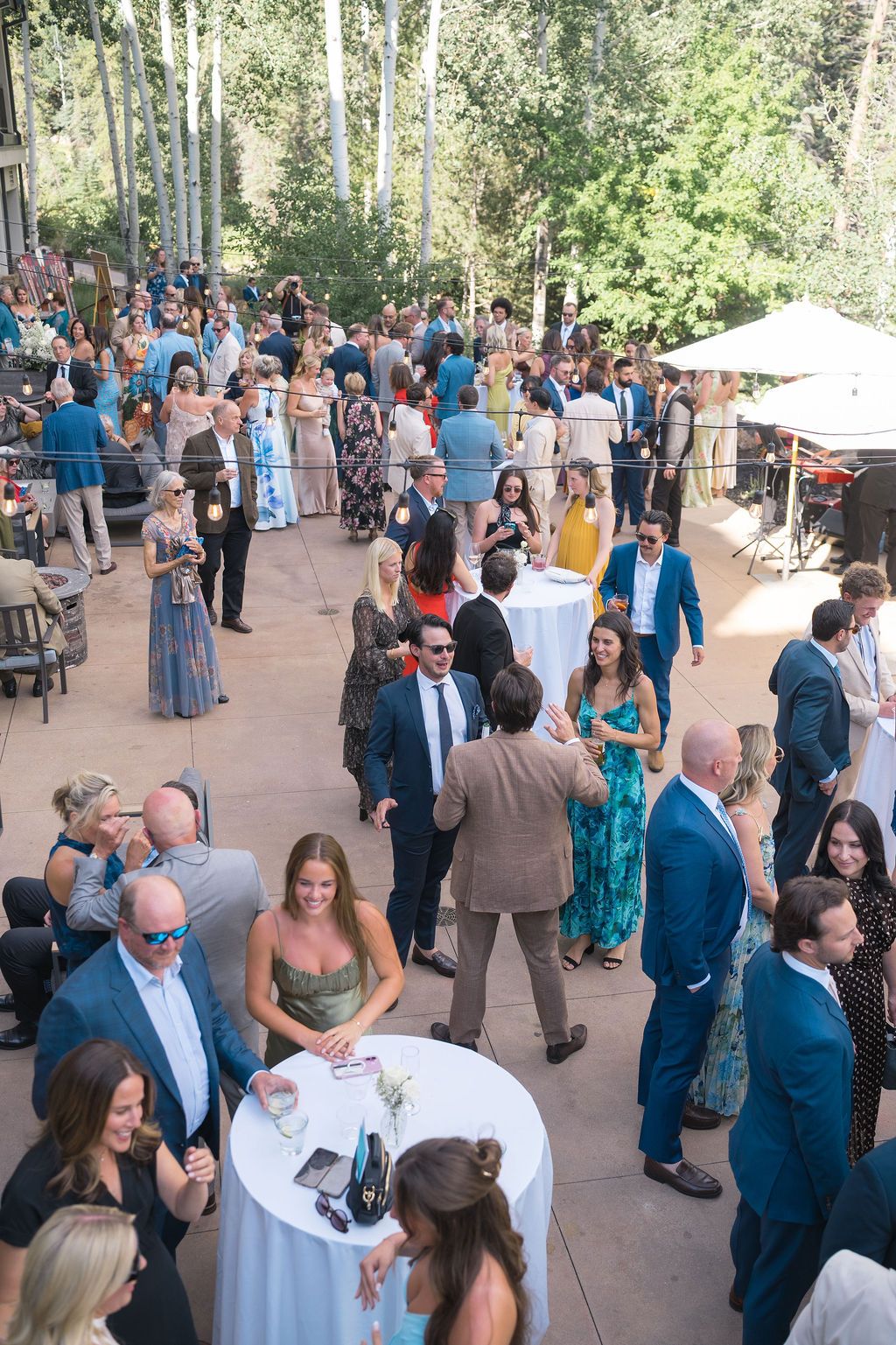 Elegant outdoor cocktail hour with guests mingling in formal attire among tall trees at a wedding reception.