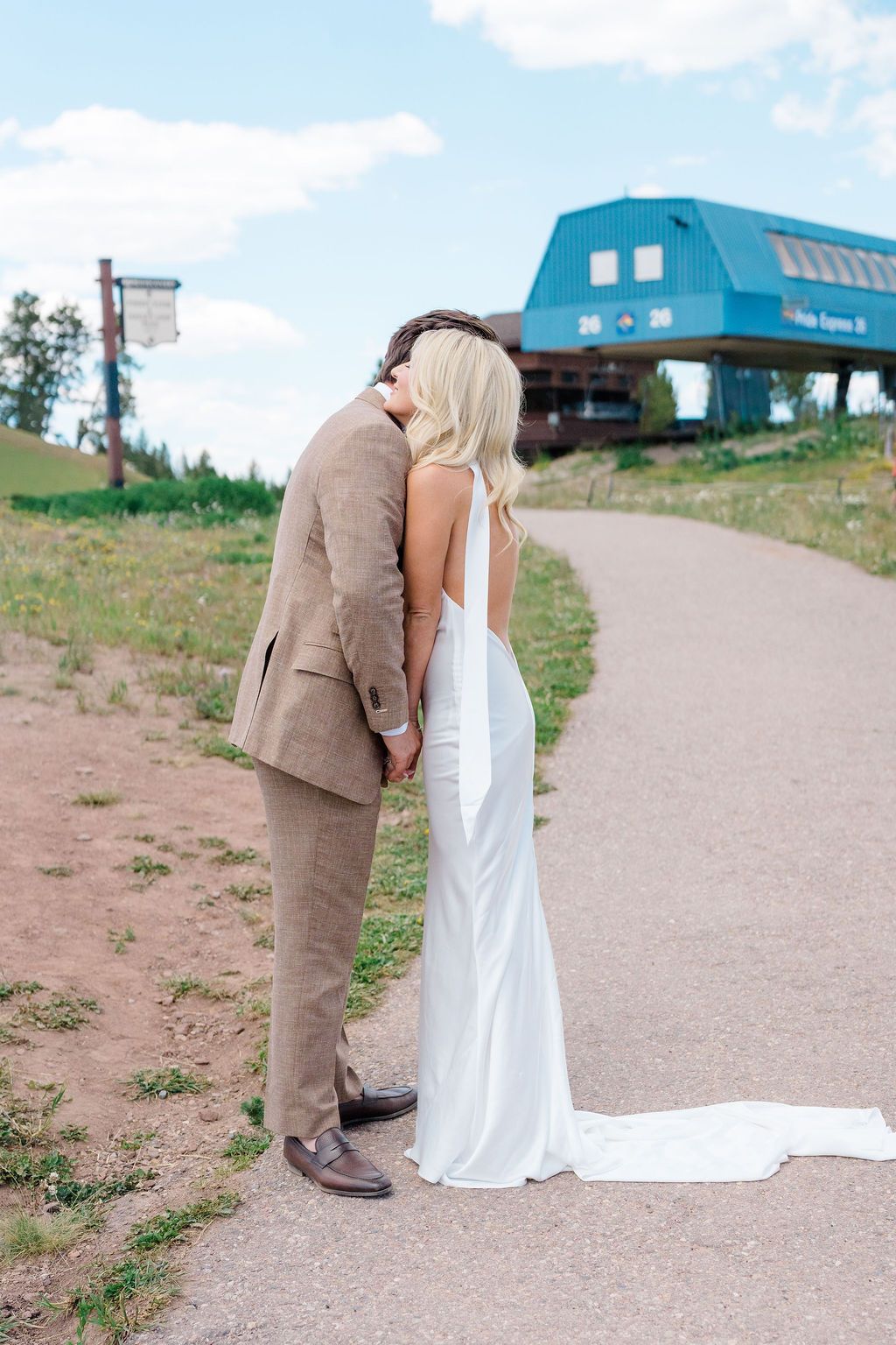 A bride in a white gown and groom in a tan suit hold hands on a scenic path near a blue mountain building.