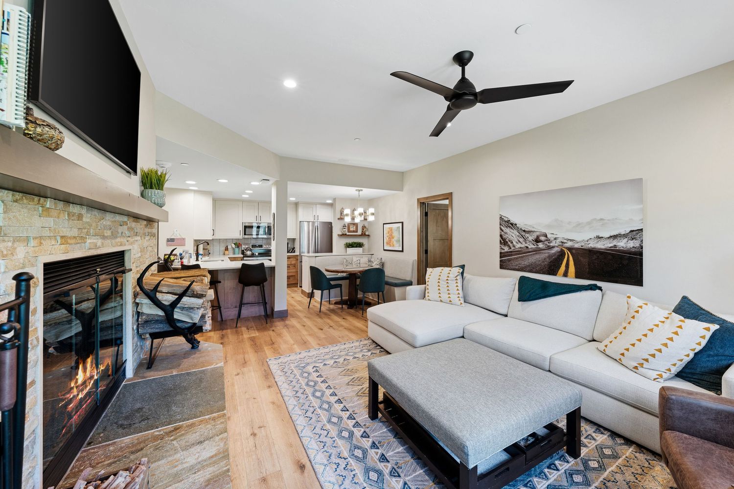 Modern living room with sectional sofa, stone fireplace, hardwood floors, and open kitchen in background.