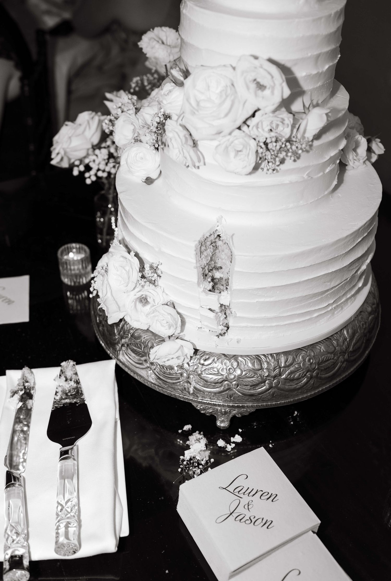 Black and white photo of a three-tier wedding cake with roses on a silver stand, beside Lauren & Jason napkins.