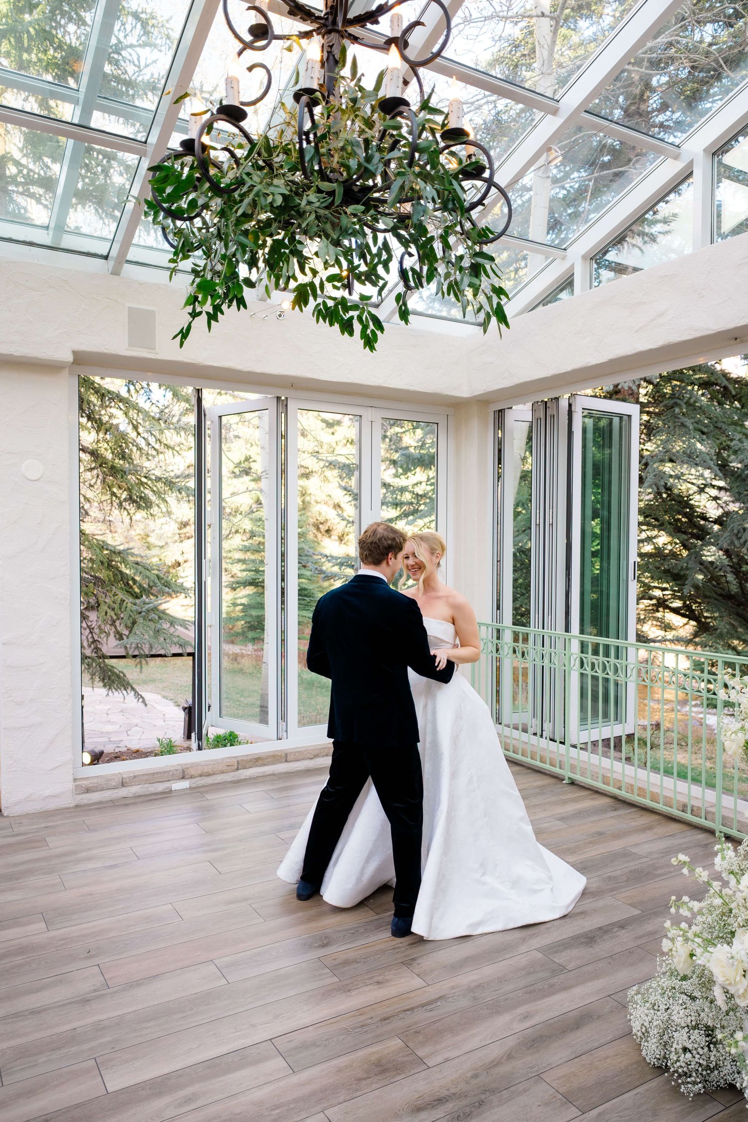 Bride and groom share first dance in elegant glass-ceiling venue with chandelier and greenery.