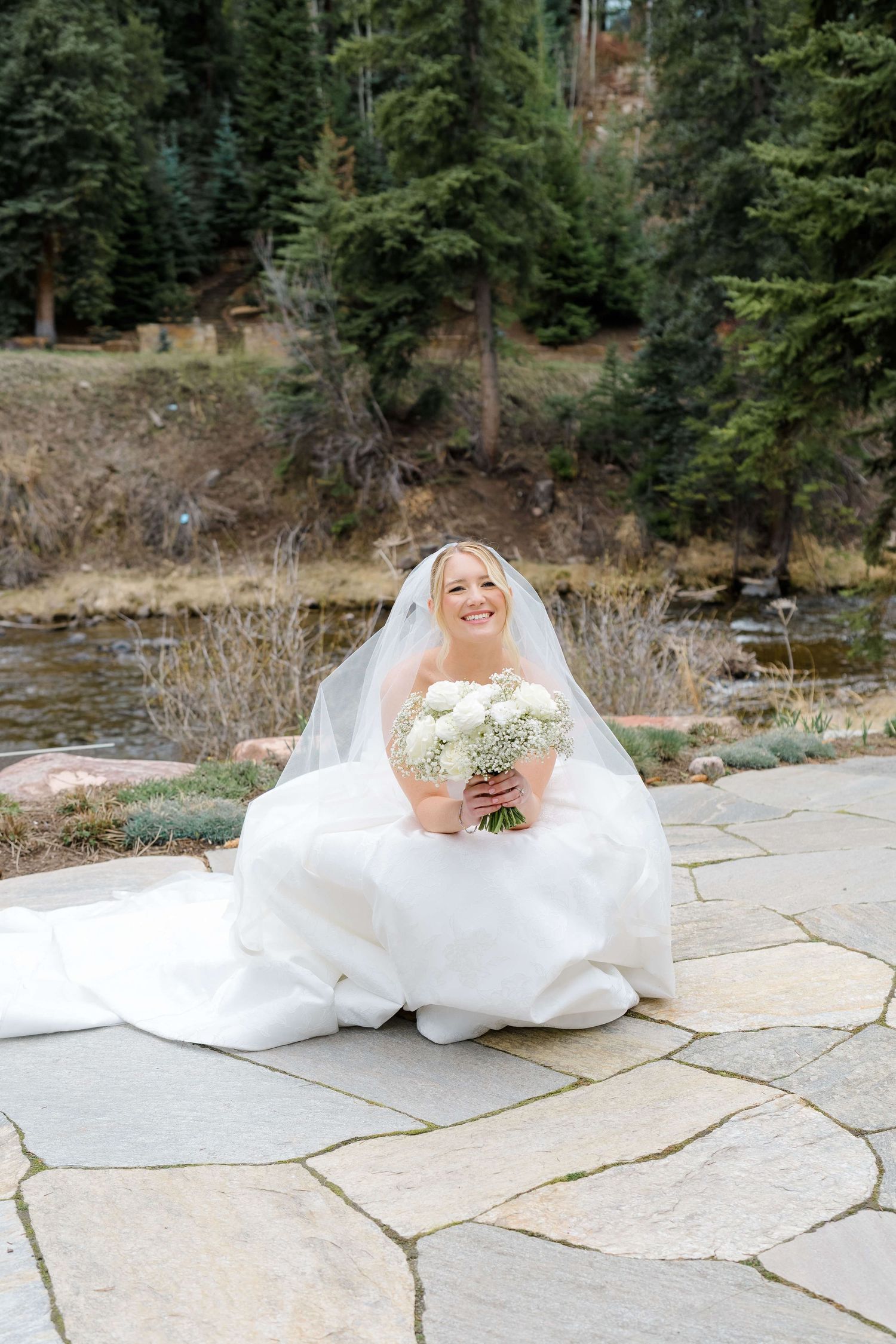 Smiling bride in white gown and veil sitting on stone patio holding white bouquet near a stream.