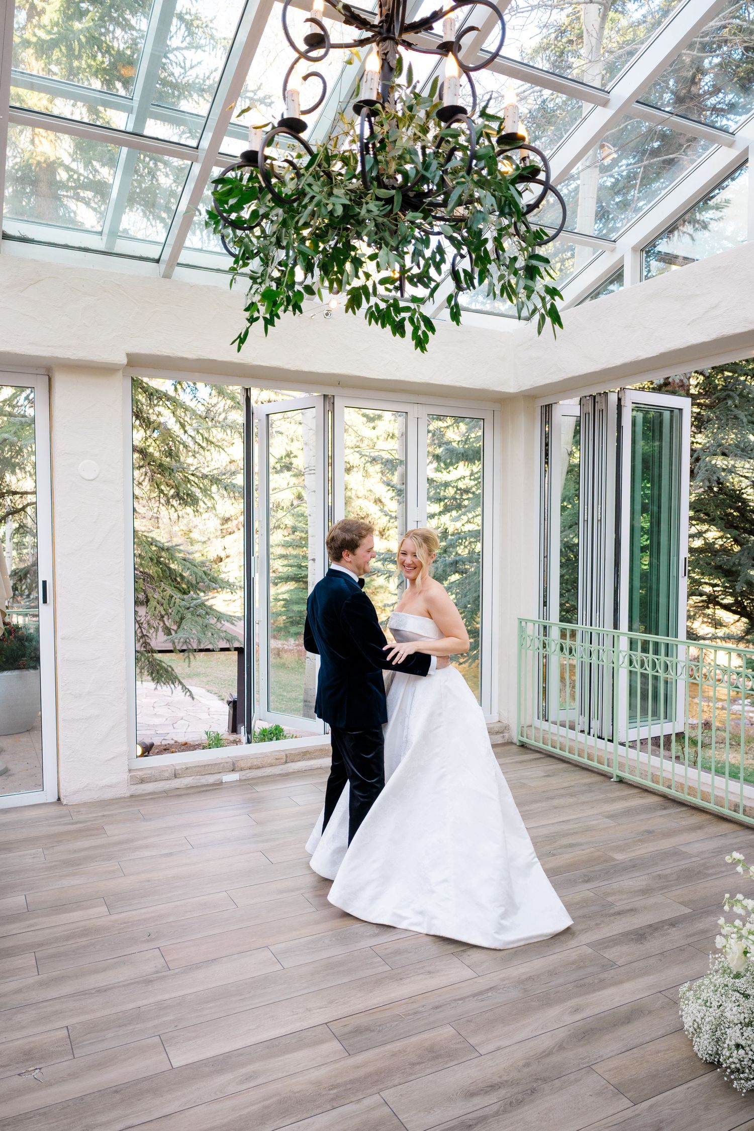 Bride and groom share first dance in elegant glass conservatory with chandelier and wood floors.