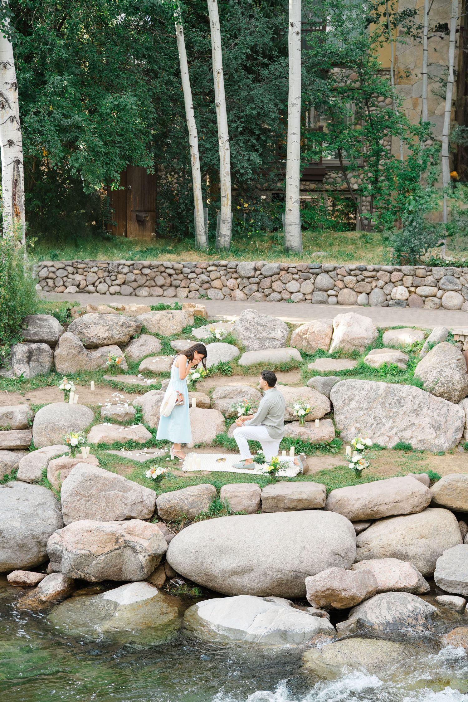 Someone relaxes on white lounge chairs by a peaceful stream surrounded by birch trees and large natural boulders.