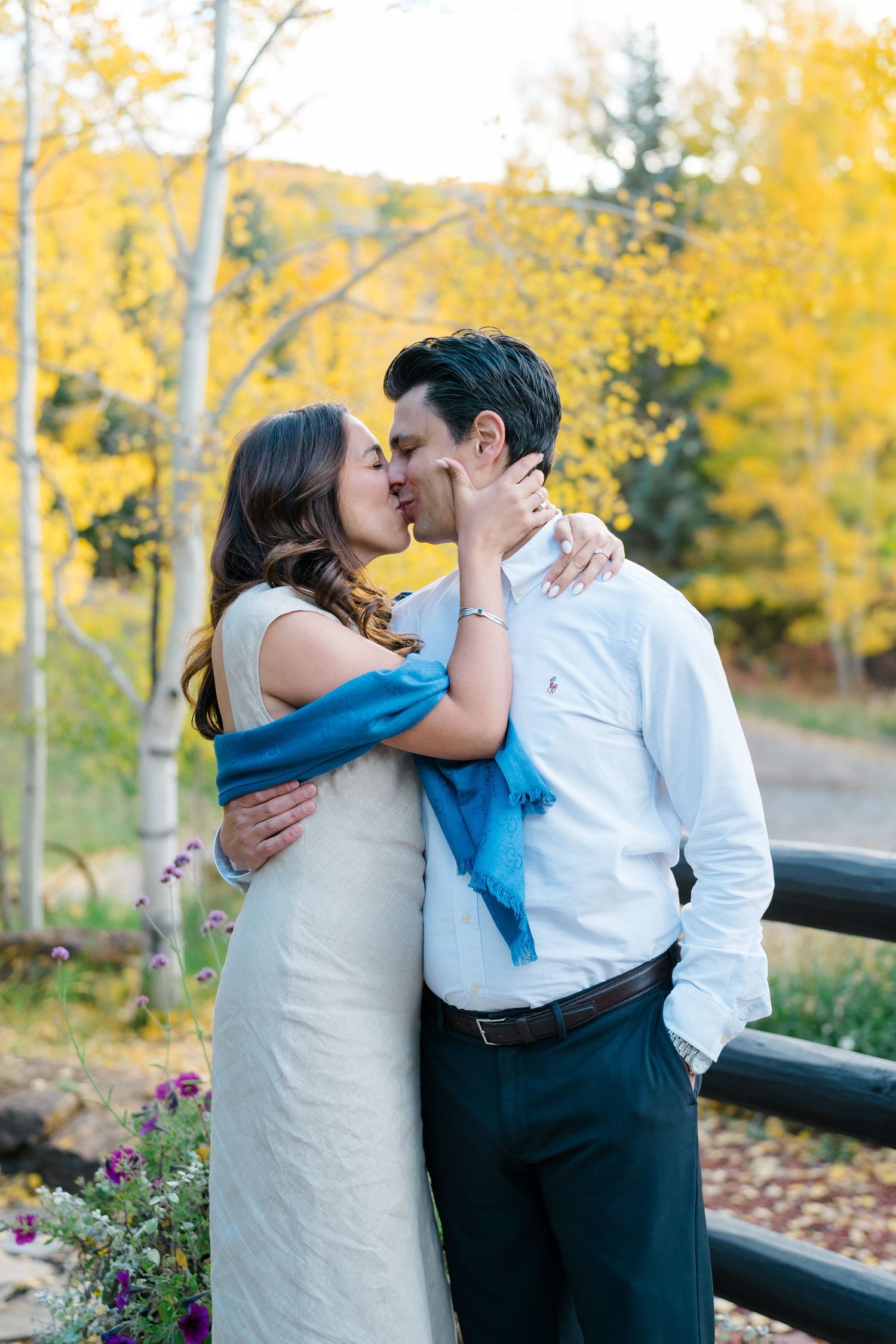 A couple shares an intimate kiss surrounded by golden autumn foliage and mountain scenery.