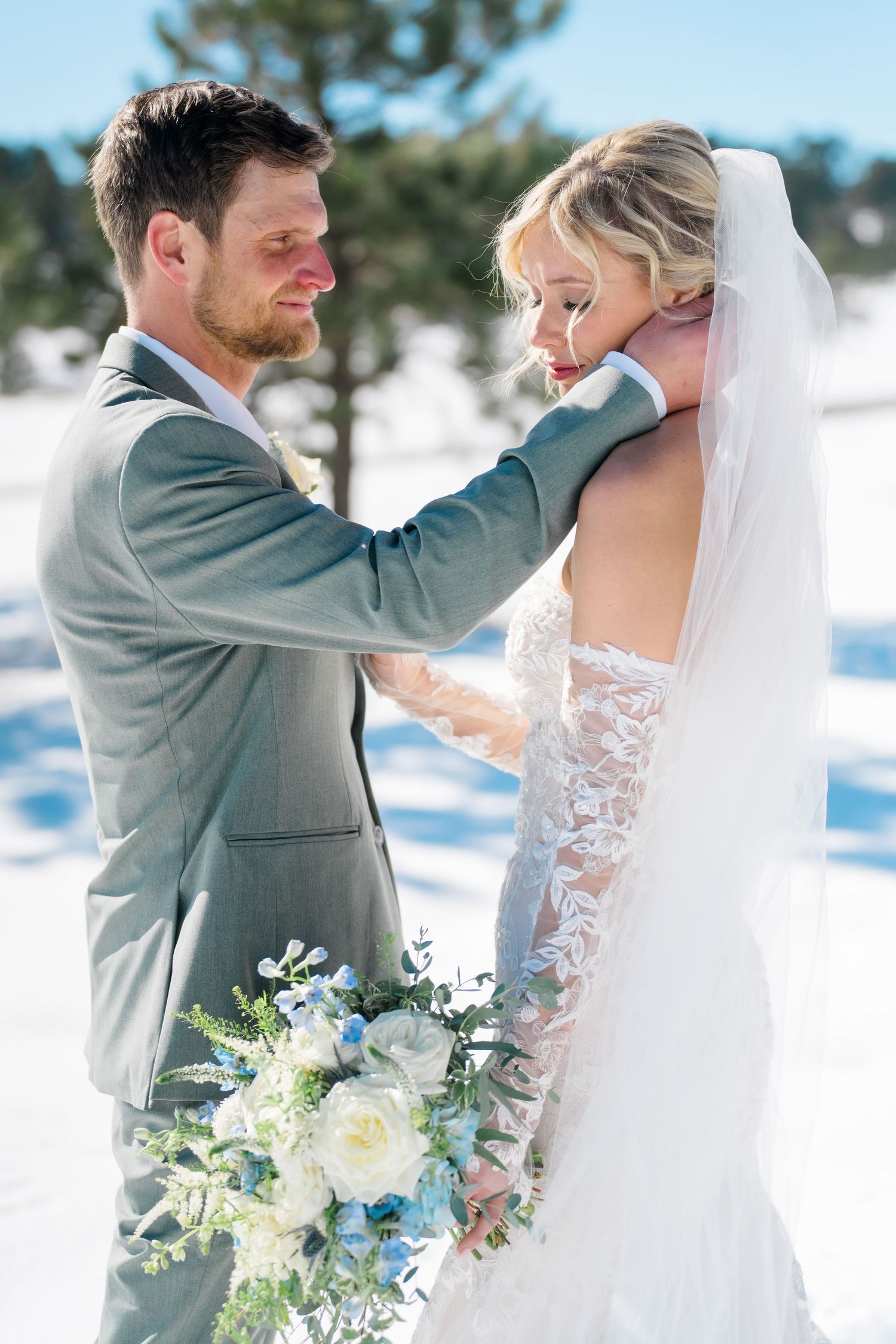 A romantic winter wedding scene with a bride and groom embracing in the snow while holding a white floral bouquet.
