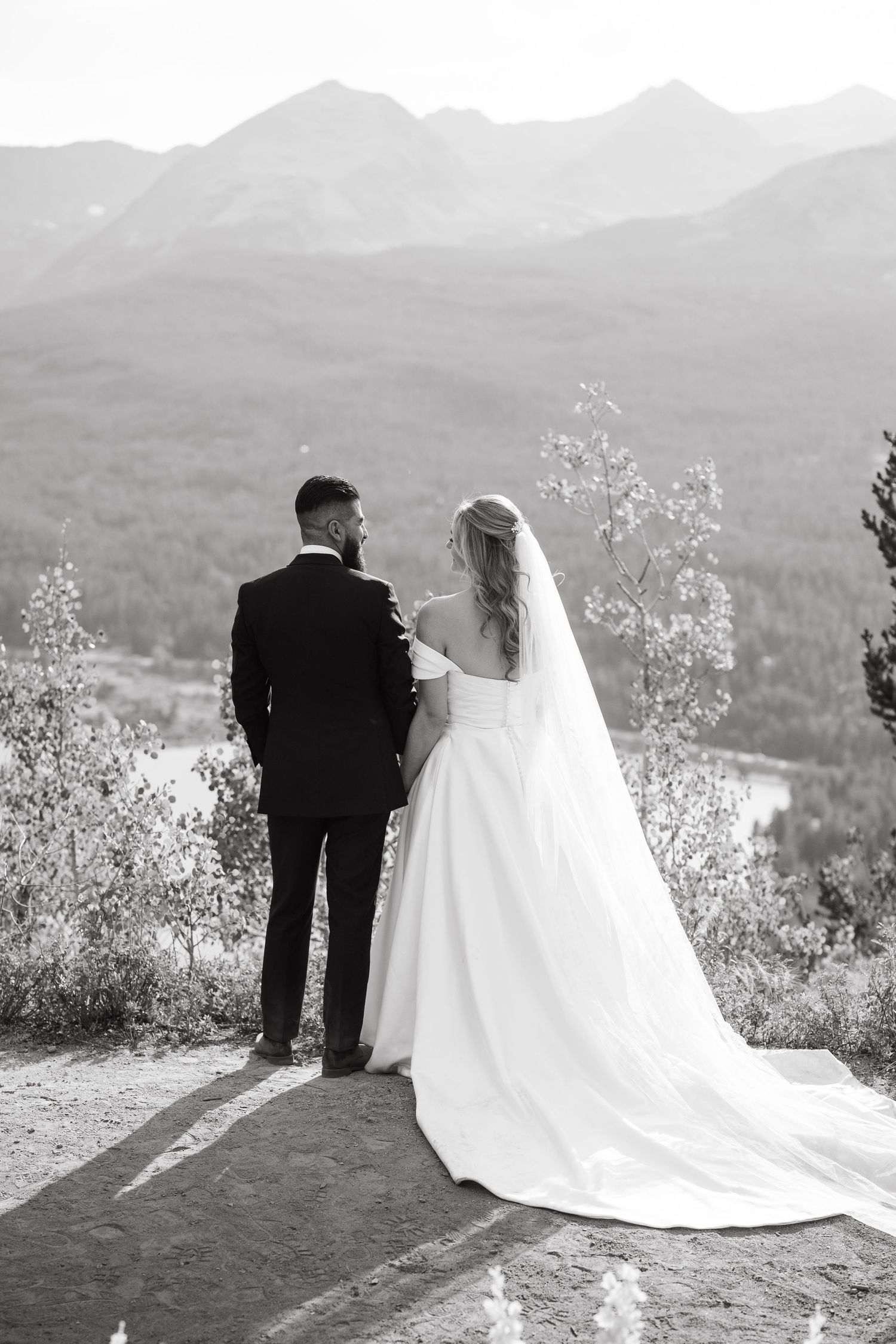 A couple in wedding attire stands on a mountaintop overlooking a scenic valley landscape in black and white.