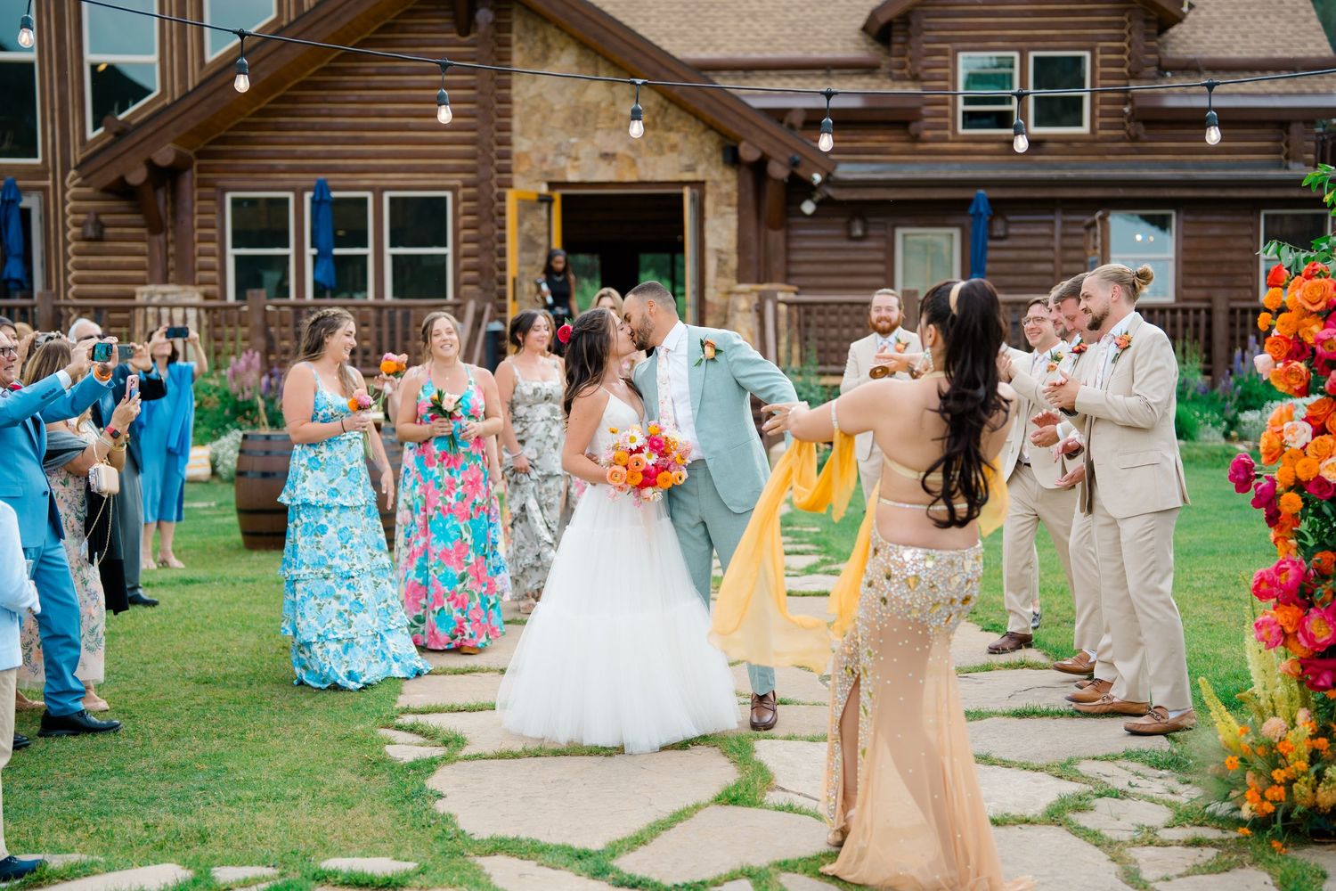Wedding guests gather on a grassy lawn outside a rustic mountain lodge during an outdoor celebration.