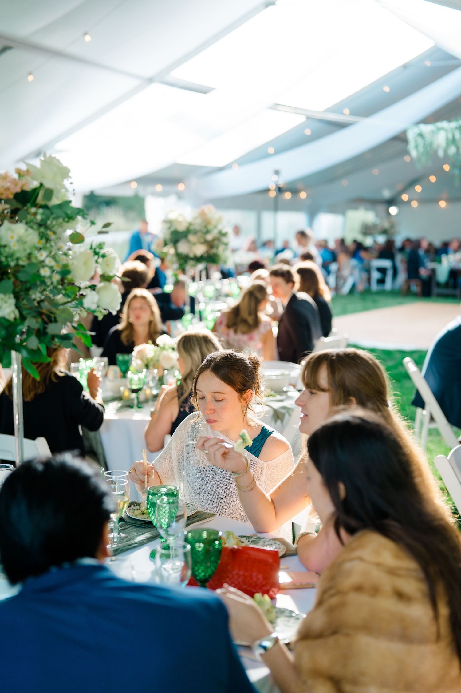 Wedding reception guests enjoy an elegant dinner under a white tent with twinkling lights and floral centerpieces.