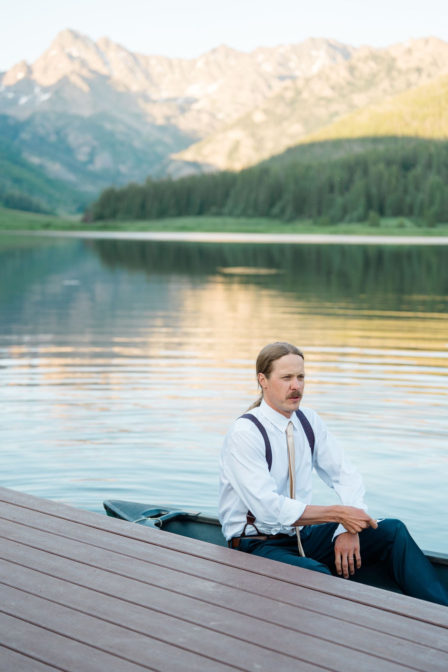 A person in white shirt and suspenders relaxes on a wooden dock by a serene mountain lake at sunset.