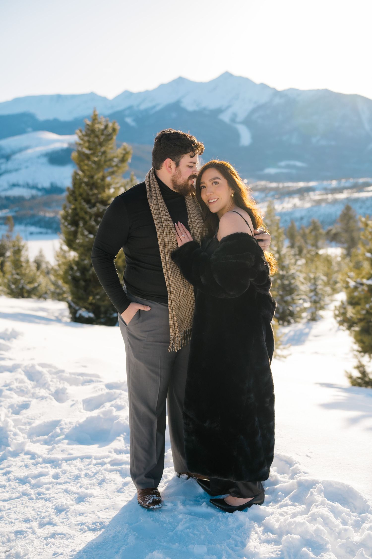 A couple shares an intimate moment in the snowy mountains with evergreen trees and mountain peaks in the background.