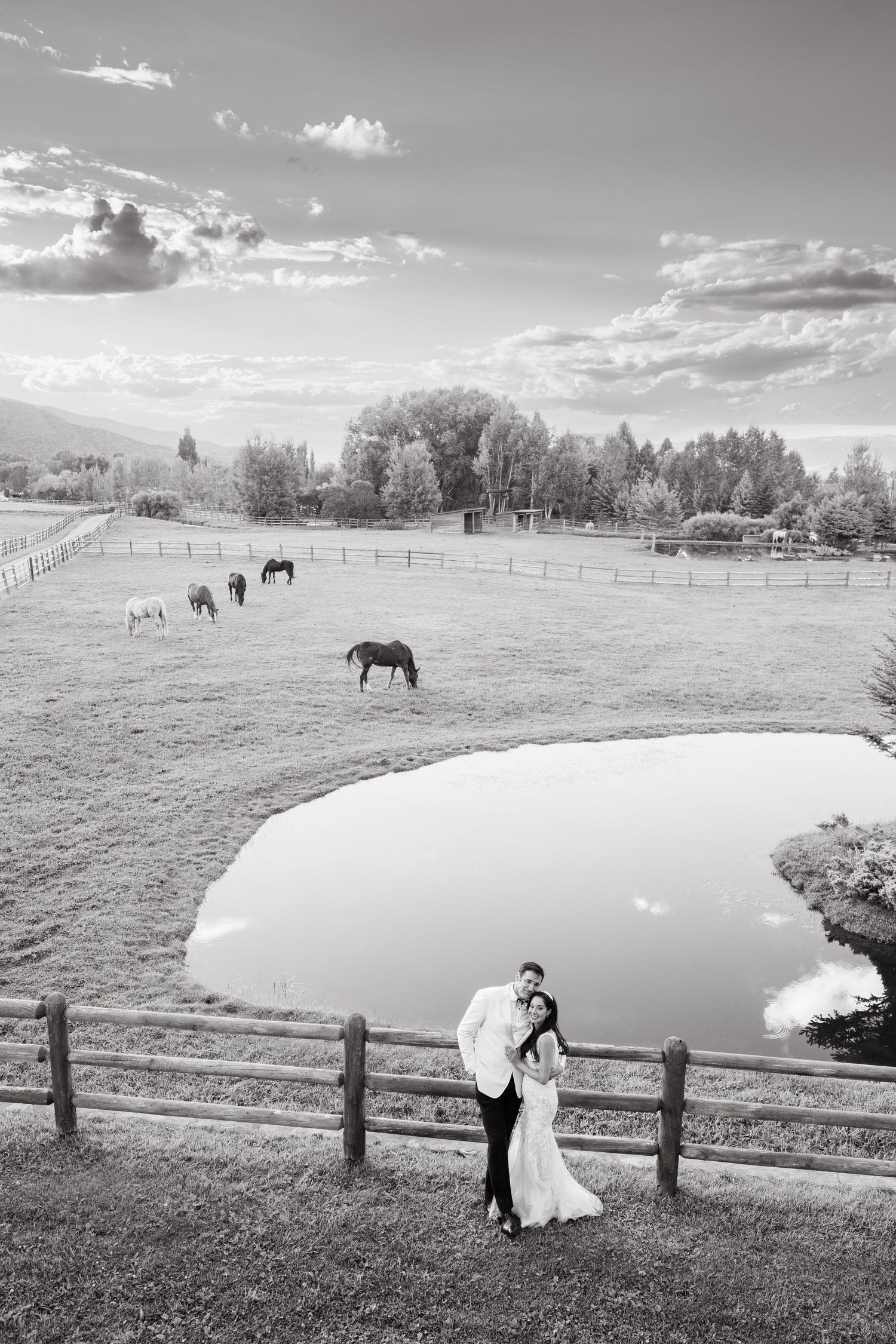 Black and white countryside scene with couple by pond and horses grazing in pastoral field under cloudy sky.
