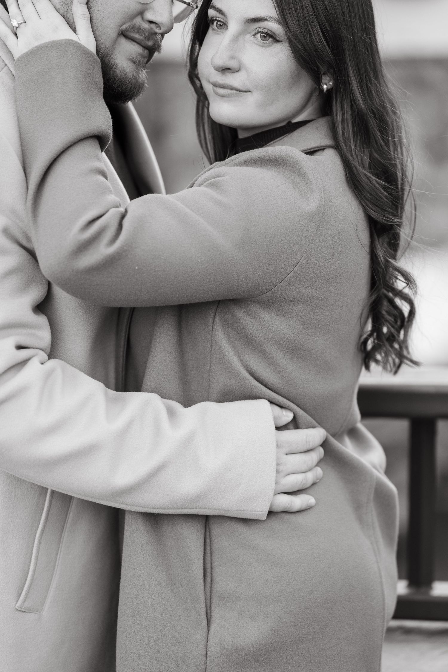 Black and white photo of a romantic couple embracing outdoors, woman in coat smiling at camera while man holds her close.