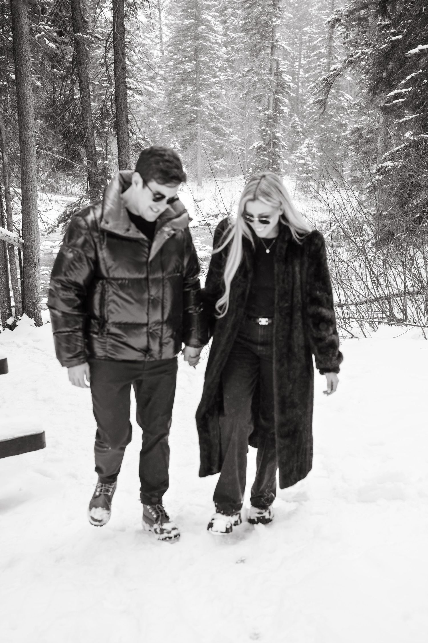 Black and white photo of a couple holding hands walking through a snowy forest trail in winter coats.