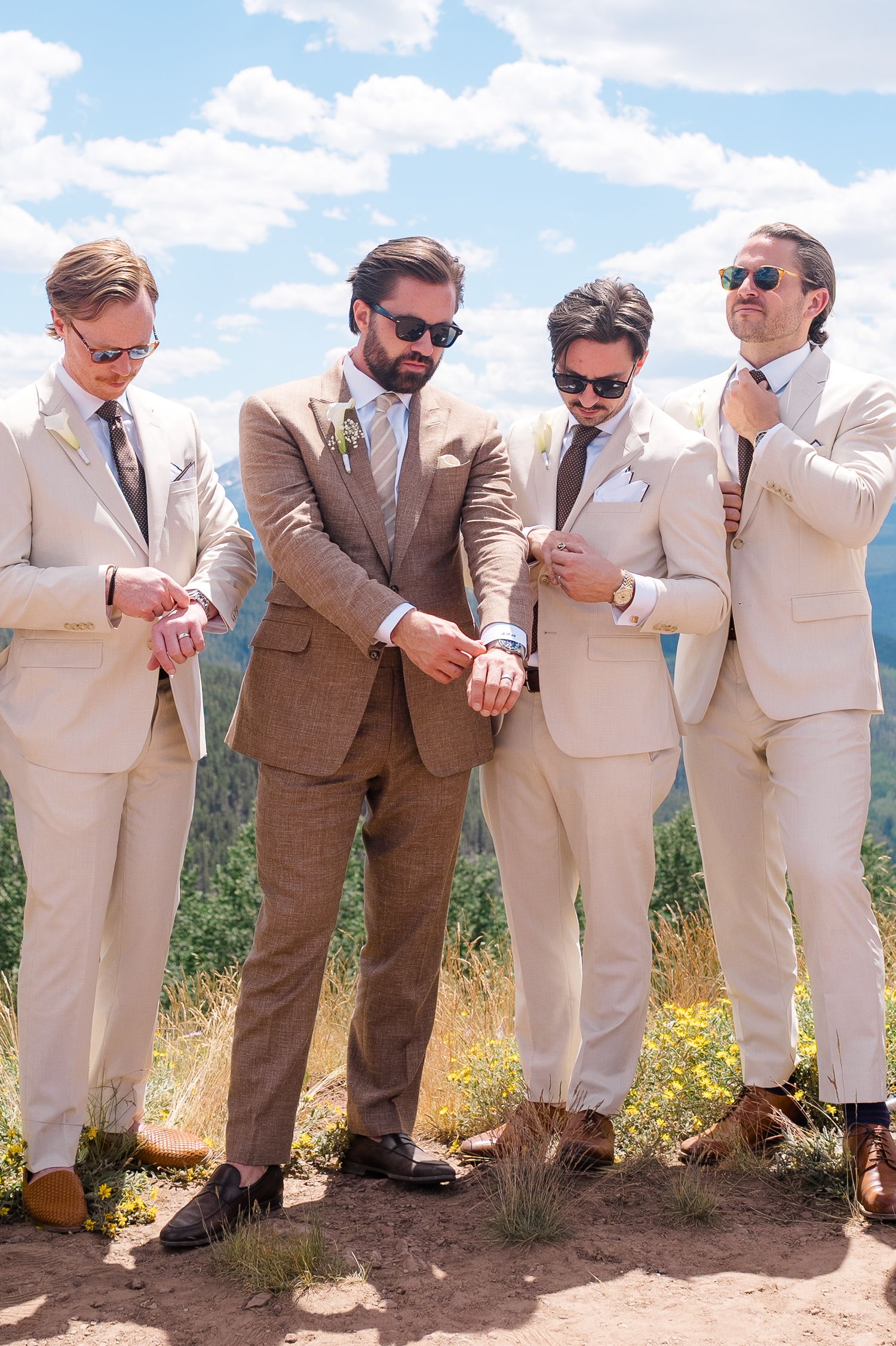 Groom in brown suit stands with groomsmen in tan suits and sunglasses outdoors on a sunny mountain wedding day.