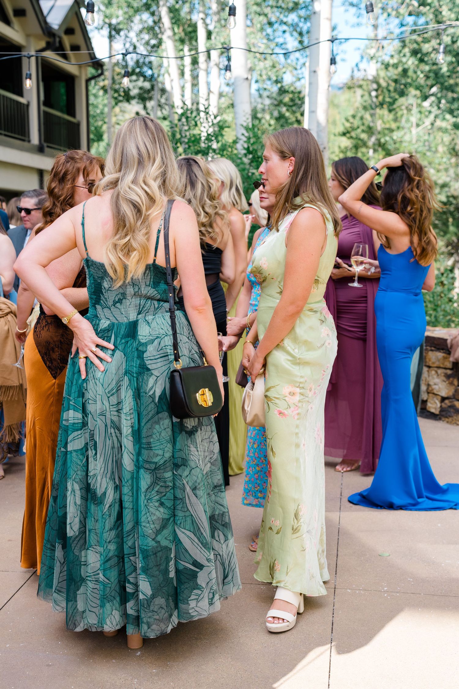 Women in elegant floral and colorful gowns socializing outdoors at a summer wedding reception.