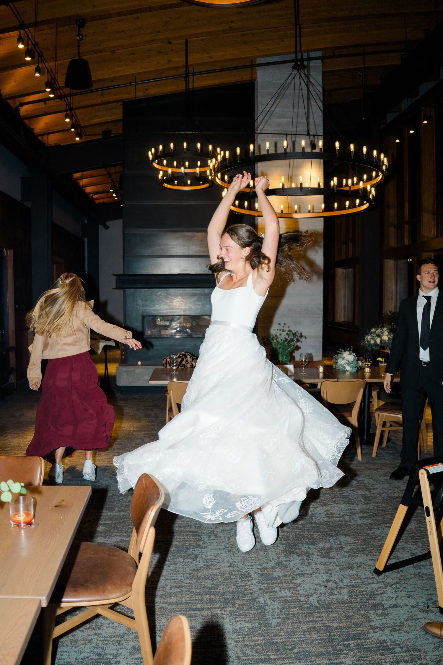 A bride in a white wedding dress and sneakers dances joyfully under circular chandeliers at a reception venue at copper mountain aerie.