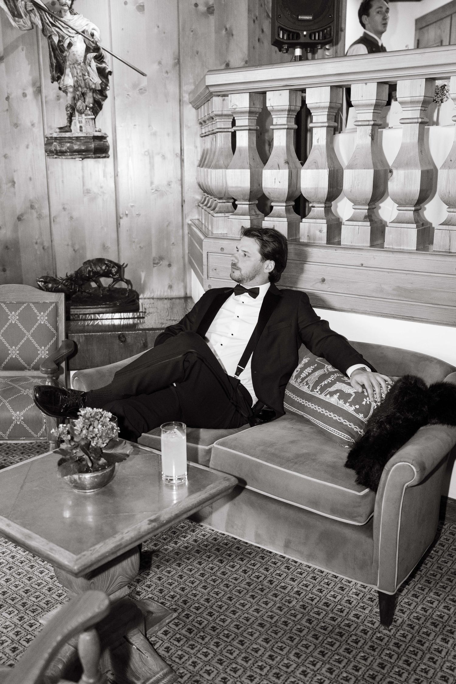 Man in tuxedo reclining on velvet sofa in elegant wood-paneled room, black and white photography.