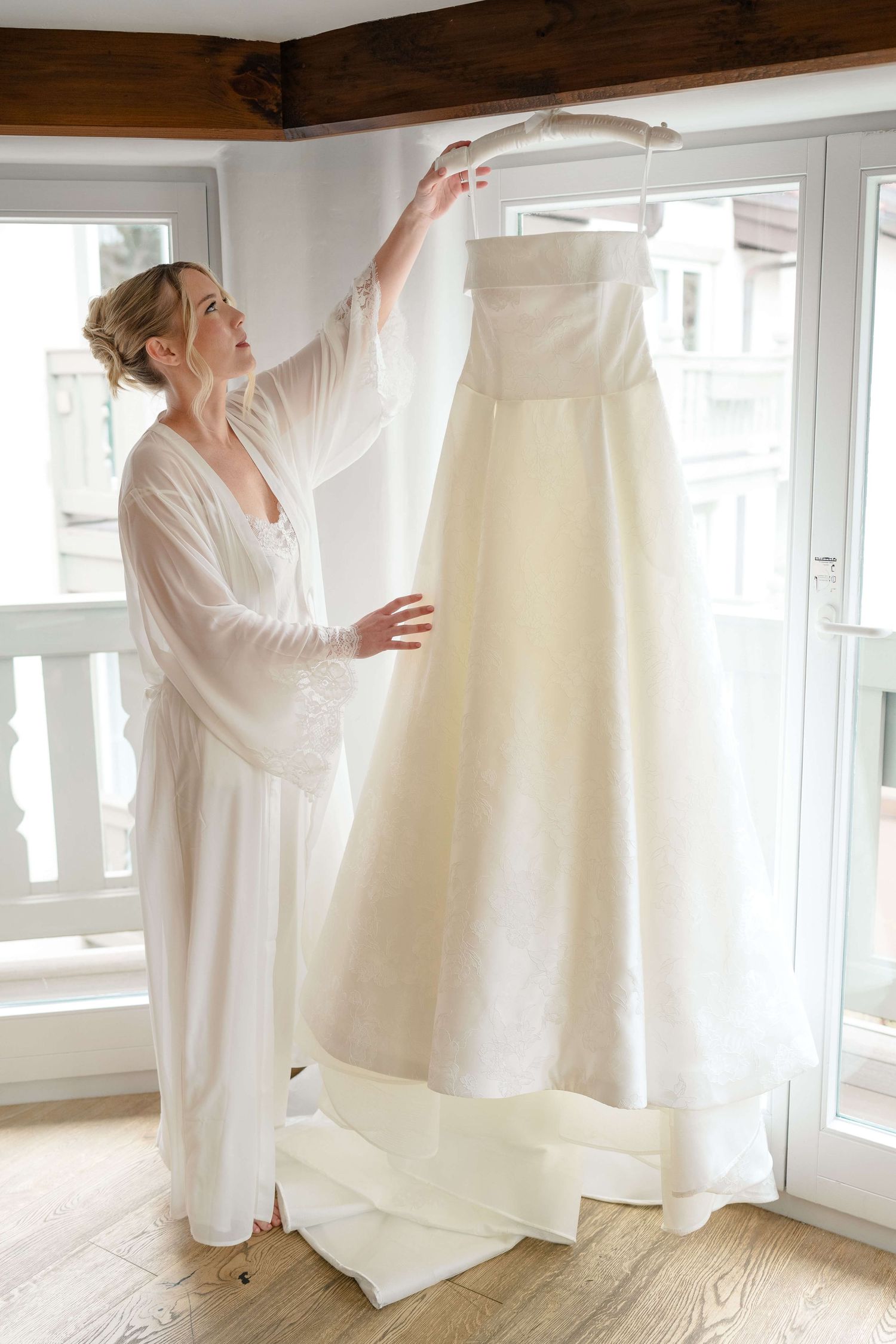 Bride in white robe admiring her elegant ivory wedding gown hanging by a bright window before the ceremony at Vail Sonnenalp Wedding.