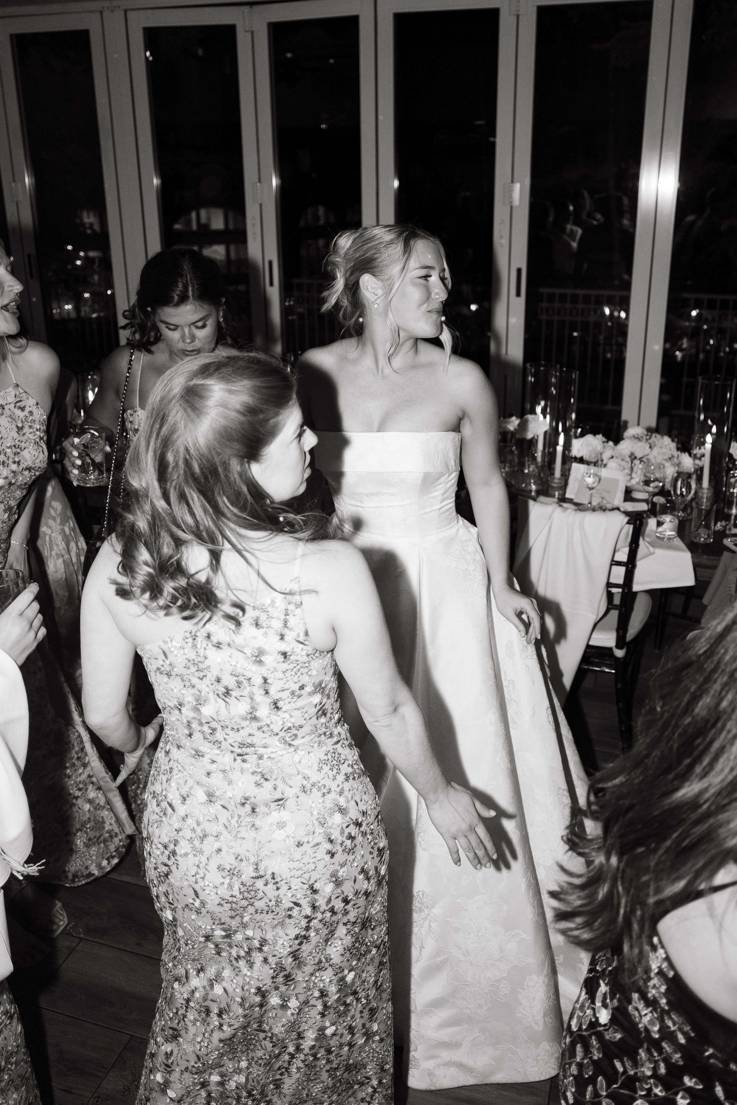 Bride in strapless white gown dancing with guests in sequin dresses at elegant wedding reception, black and white photo.
