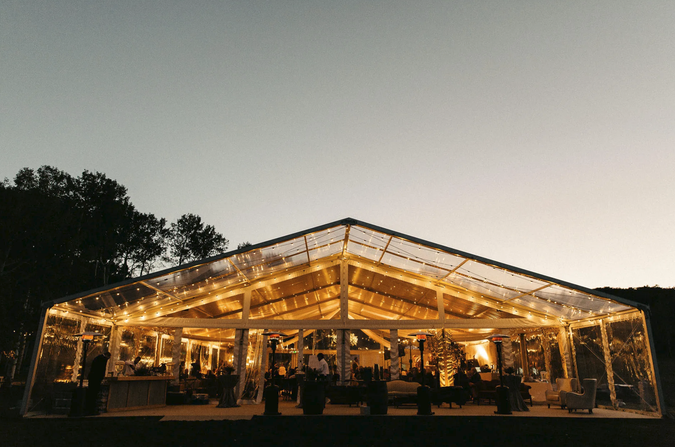 Elegant outdoor event tent glowing with warm string lights at dusk, surrounded by trees and guests.