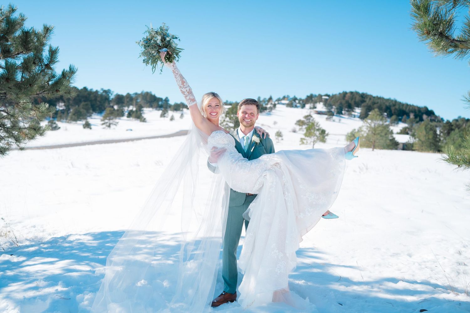 Groom carries bride in snowy winter wedding landscape with pine trees and blue sky in Colorado.