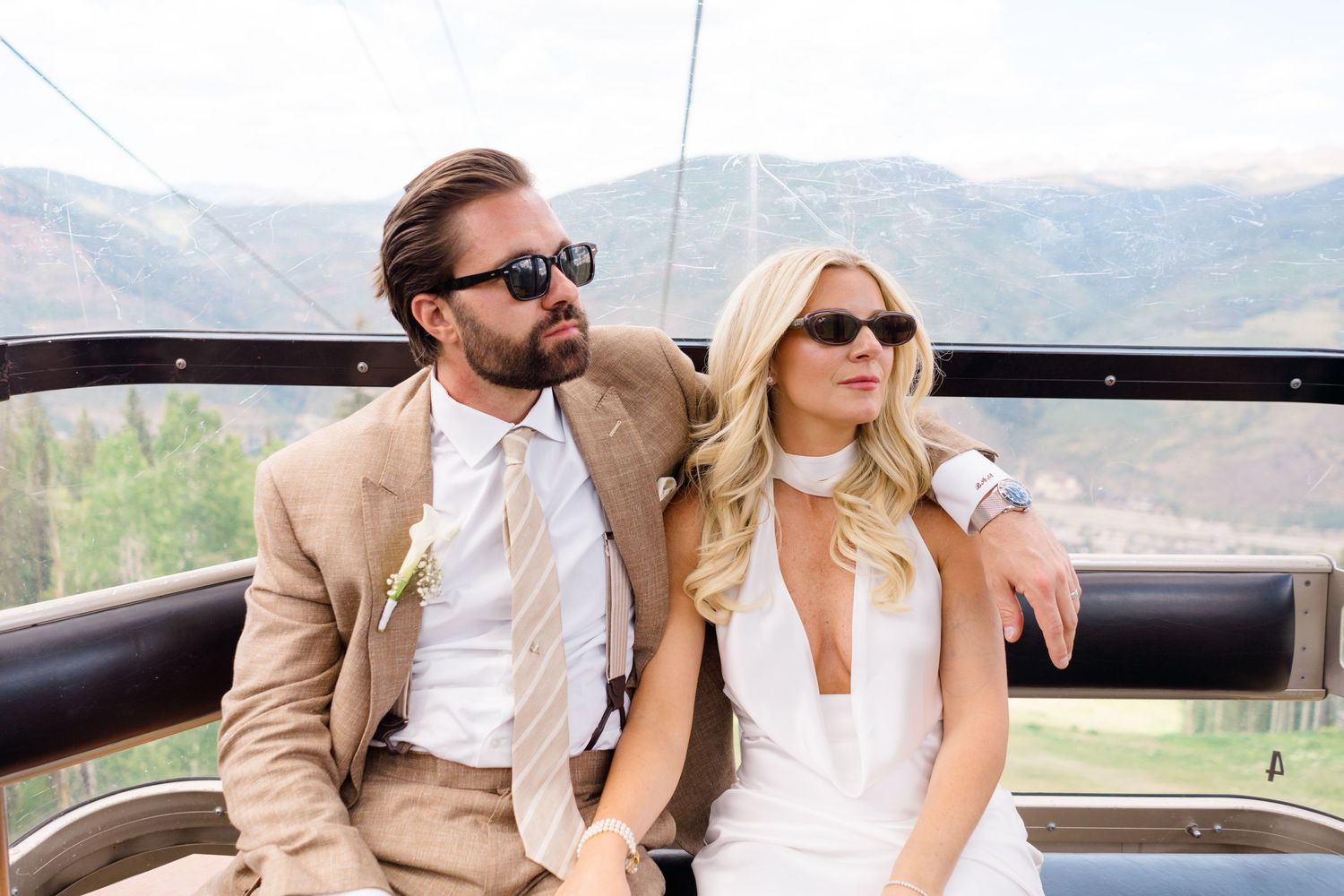Stylish couple in sunglasses sitting on a gondola lift, man in tan suit, woman in white dress, mountains in background.
