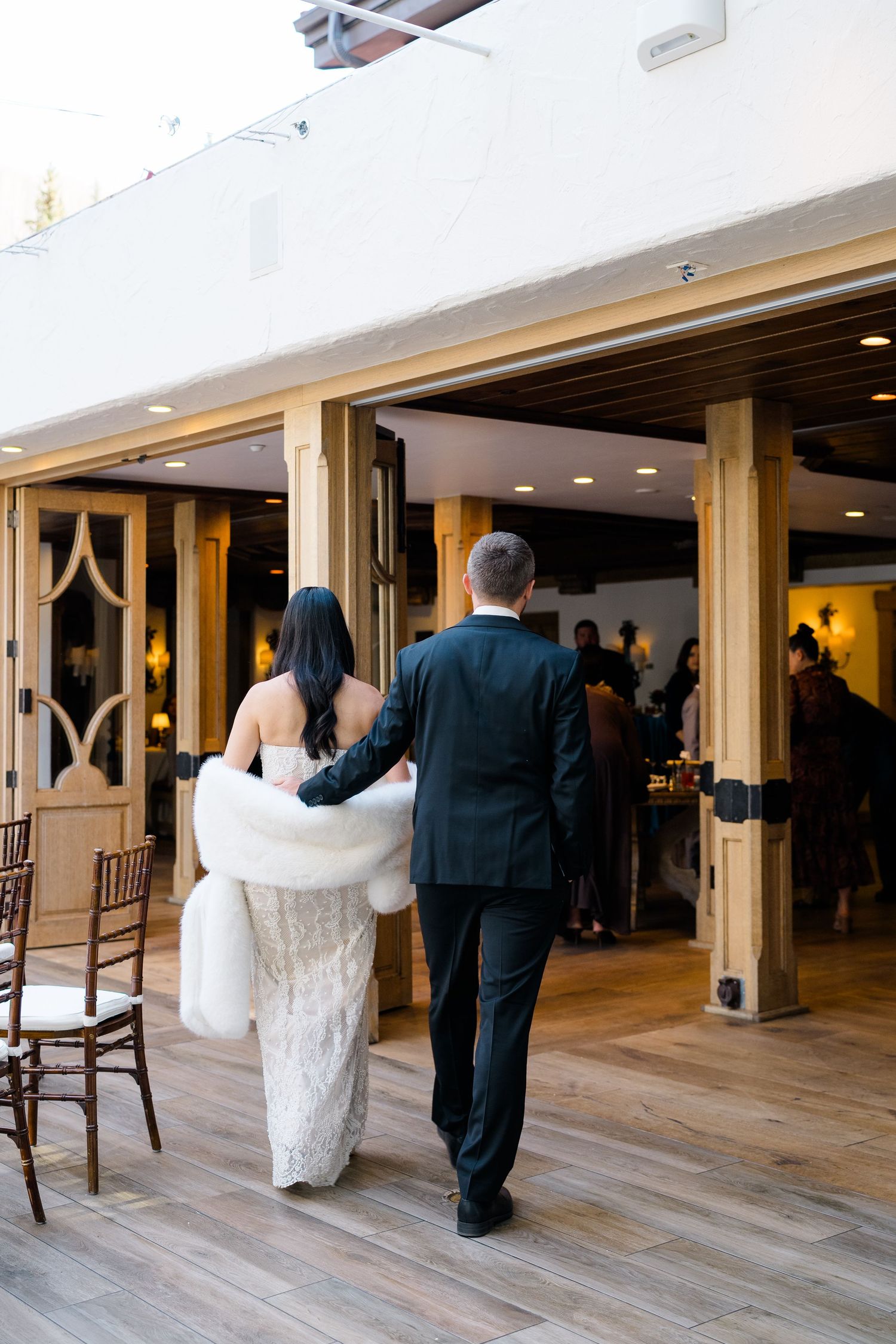 Bride in white lace gown with fur stole walks arm-in-arm with groom in black suit into wedding reception venue.