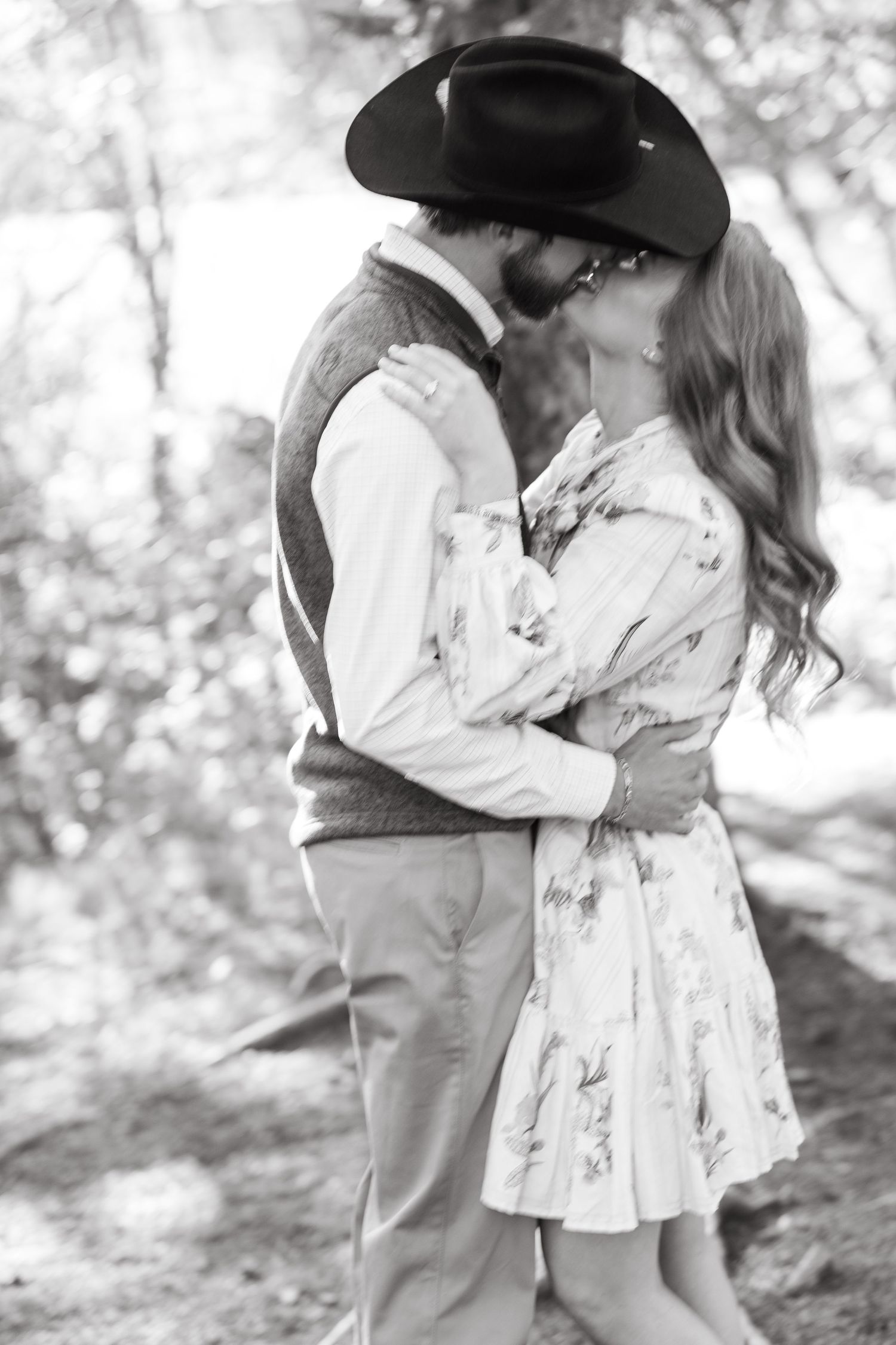 Black and white photo of a couple kissing outdoors, man wearing cowboy hat, woman in floral dress embracing romantically.